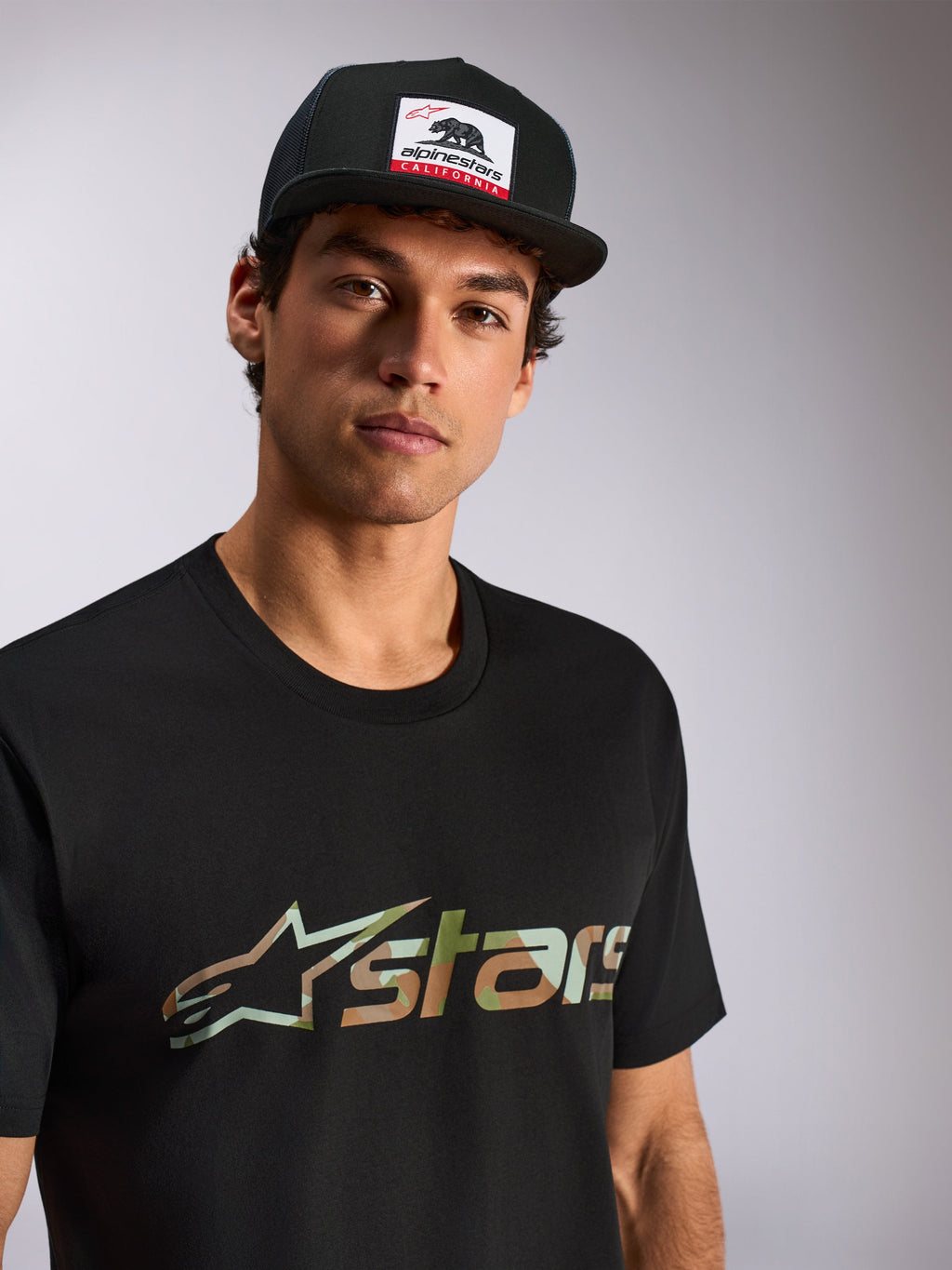 Alpinestars Verdure Camo CSF Tee, kurzärmeliges T-Shirt, Schwarz, Astars-Logo mit Camo-Füllung, getragen von einem männlichen Model mit schwarzer Alpinestars California Trucker-Kappe, lässige Motorsport-Streetwear