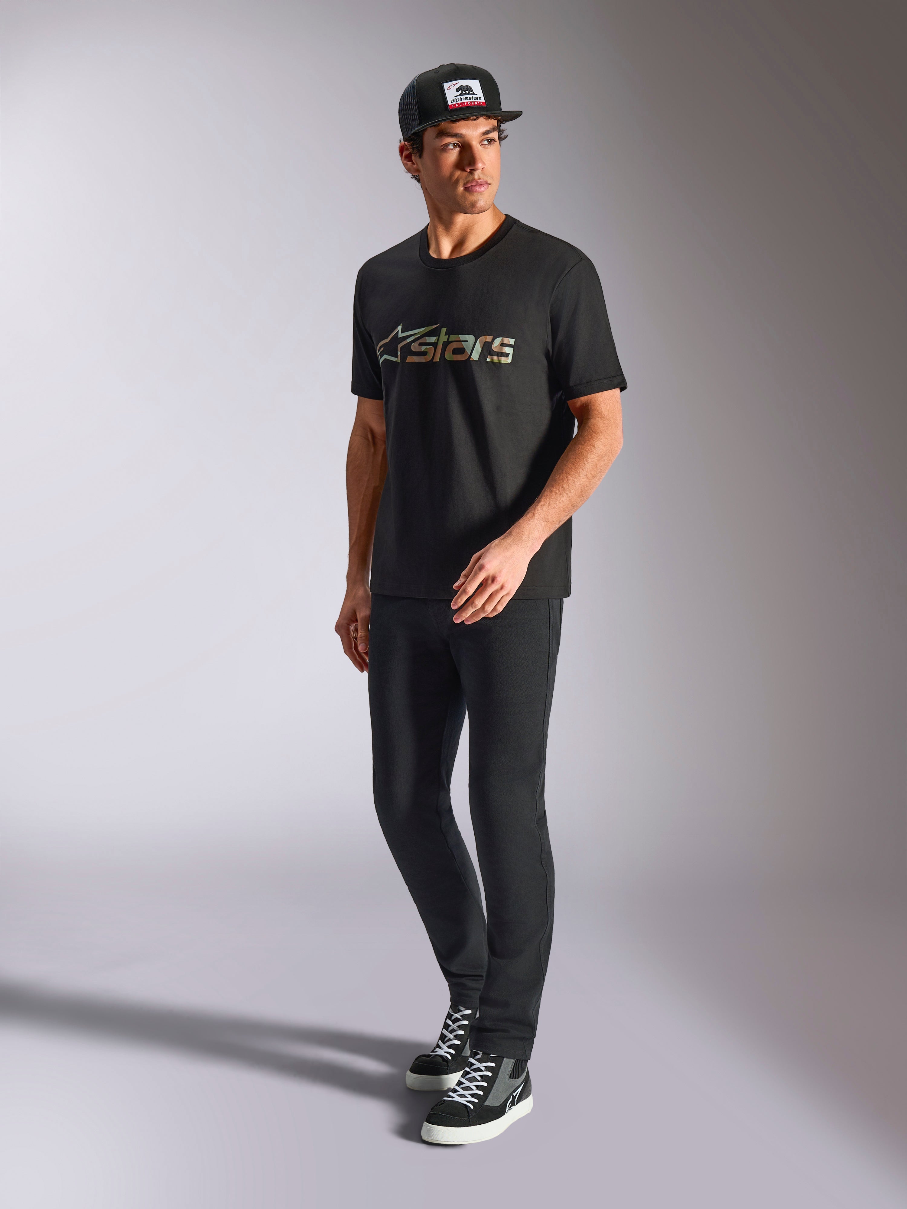 Alpinestars Verdure Camo CSF Tee, Kurzärmeliges T-Shirt, Schwarz, mit einem Astars-Logo mit Camouflage-Füllung auf der Brust, getragen von einem Model mit schwarzer Kappe und Hose, lässige Motorsport-Streetwear