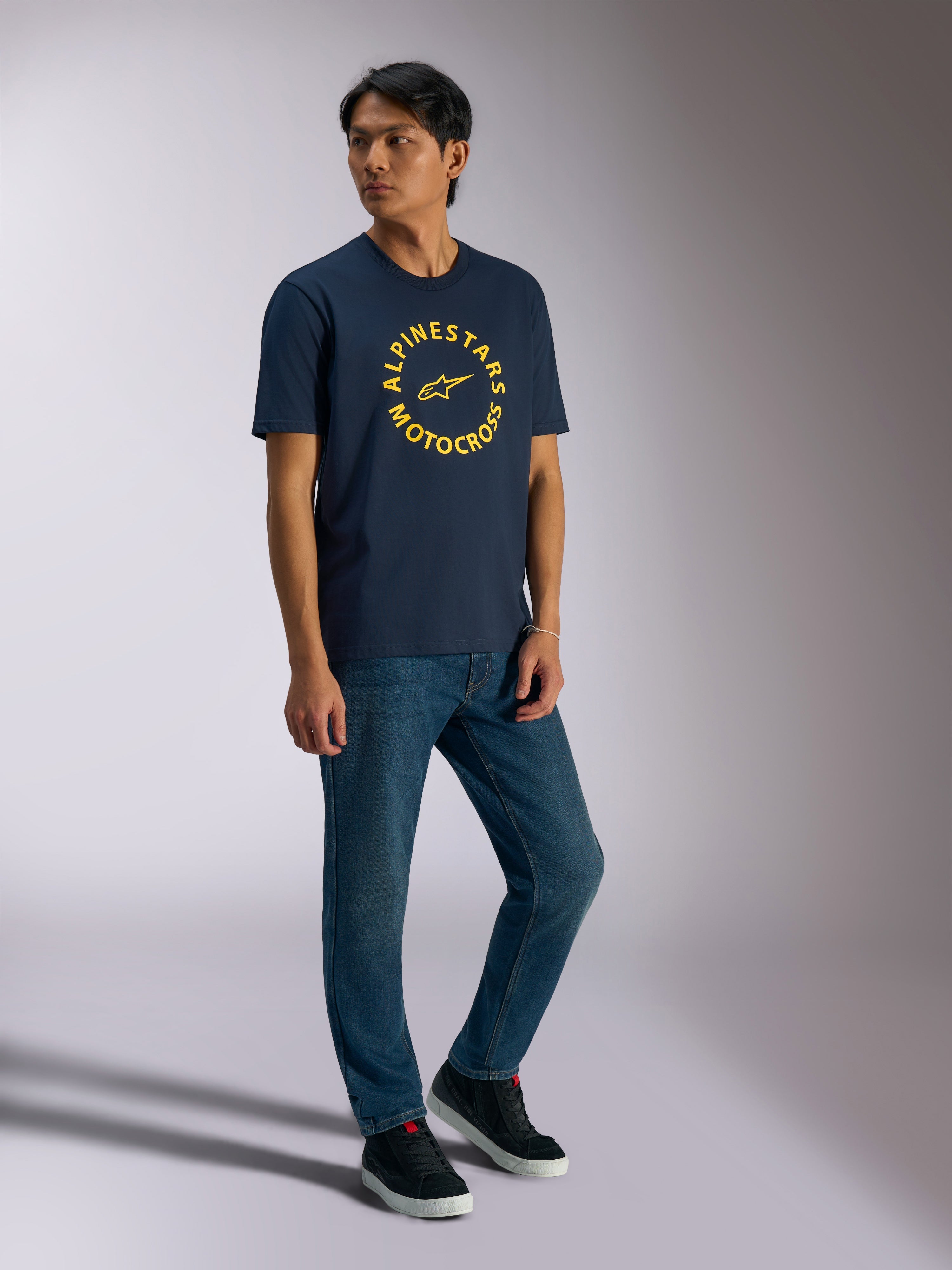 Alpinestars MX CSF Tee, Kurzarm-T-Shirt, Navy, gelbe kreisförmige Alpinestars Motocross-Grafik, getragen von einem Model mit blauen Jeans und schwarzen Sneakern, lässige Motorsport-Streetwear