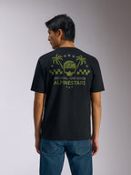 Alpinestars Order CSF Tee, Kurzarm-T-Shirt, Schwarz, Rückansicht, olivgrüne Grafik mit Palmen und Helm-Logo, Text „ONE GOAL. ONE VISION. ALPINESTARS“, kombiniert mit blauen Jeans, lässige Motorsport-Streetwear