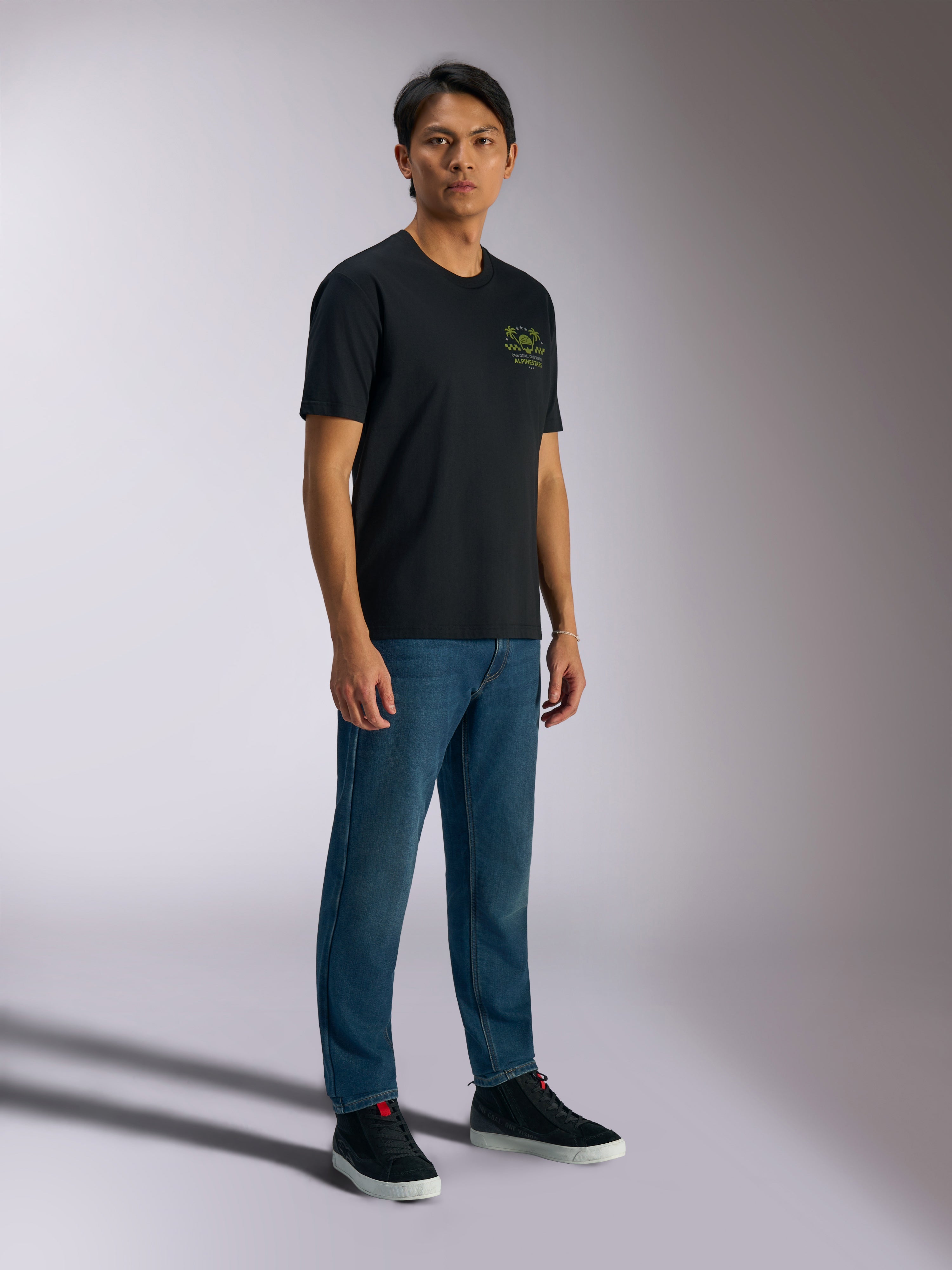 Alpinestars Order CSF Tee, kurzärmeliges T-Shirt, Schwarz, mit grünen Helm- und Palmenlogos auf der Brust, lässige Motorsport-Streetwear, kombiniert mit blauen Jeans und schwarzen Sneakern