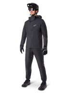 Alpinestars A-Dura Elite Waterproof Jacket in Black, worn by a male rider with matching black MTB pants, gloves, and helmet. The jacket features a technical waterproof design with a white Astars logo on the chest and a high protective collar.