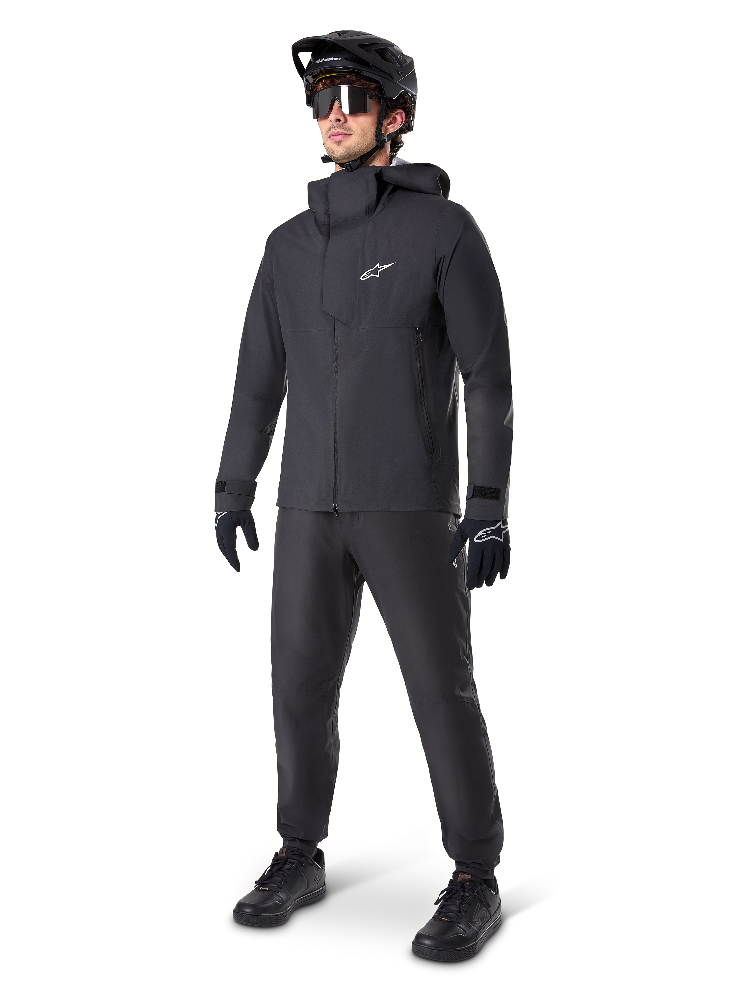 Alpinestars A-Dura Elite Waterproof Jacket in Black, worn by a male rider with matching black MTB pants, gloves, and helmet. The jacket features a technical waterproof design with a white Astars logo on the chest and a high protective collar.