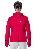 Alpinestars A-Dura Elite Wasserdichte Jacke, Mountainbike-Regenjacke, Rot Fluo, hoher Kragen mit weißem Alpinestars-Logo, schwarze Reißverschluss-Details, getragen mit passenden Handschuhen und schwarzem Fahrradhelm, technische wasserdichte Fahrradausrüstung