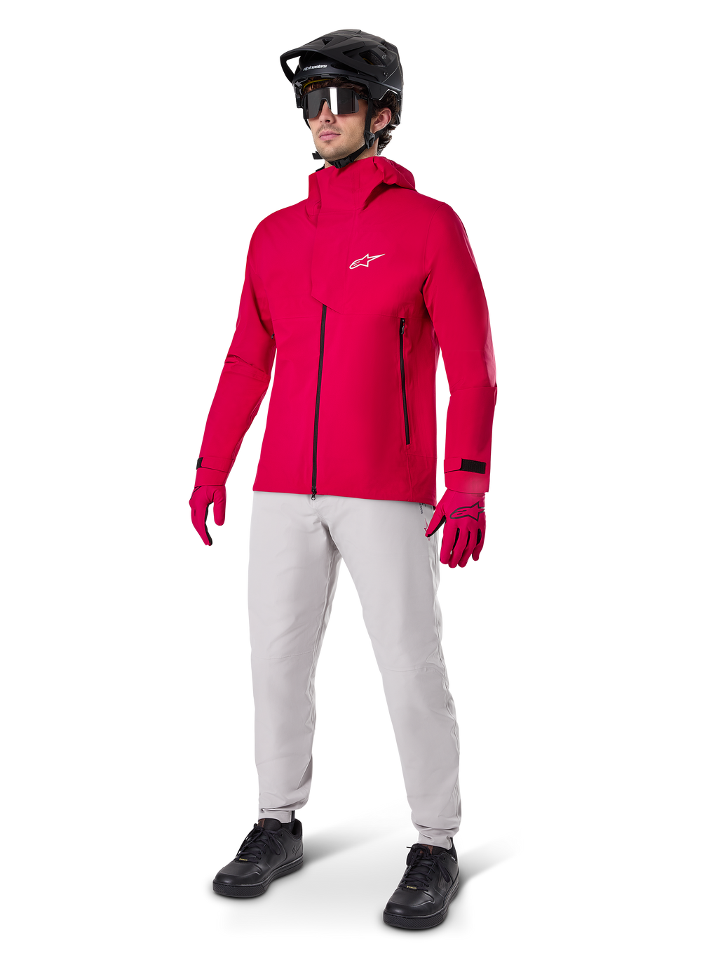 Männlicher Radfahrer in der Alpinestars A-Dura Elite wasserdichten Jacke in Red Fluo, kombiniert mit hellgrauer technischer Hose, roten Handschuhen, schwarzem MTB-Helm mit Brille und schwarzen Schuhen, komplettes Hochleistungs-Outfit für Mountainbiking
