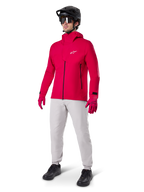 Männlicher Radfahrer in der Alpinestars A-Dura Elite wasserdichten Jacke in Red Fluo, kombiniert mit hellgrauer technischer Hose, roten Handschuhen, schwarzem MTB-Helm mit Brille und schwarzen Schuhen, komplettes Hochleistungs-Outfit für Mountainbiking