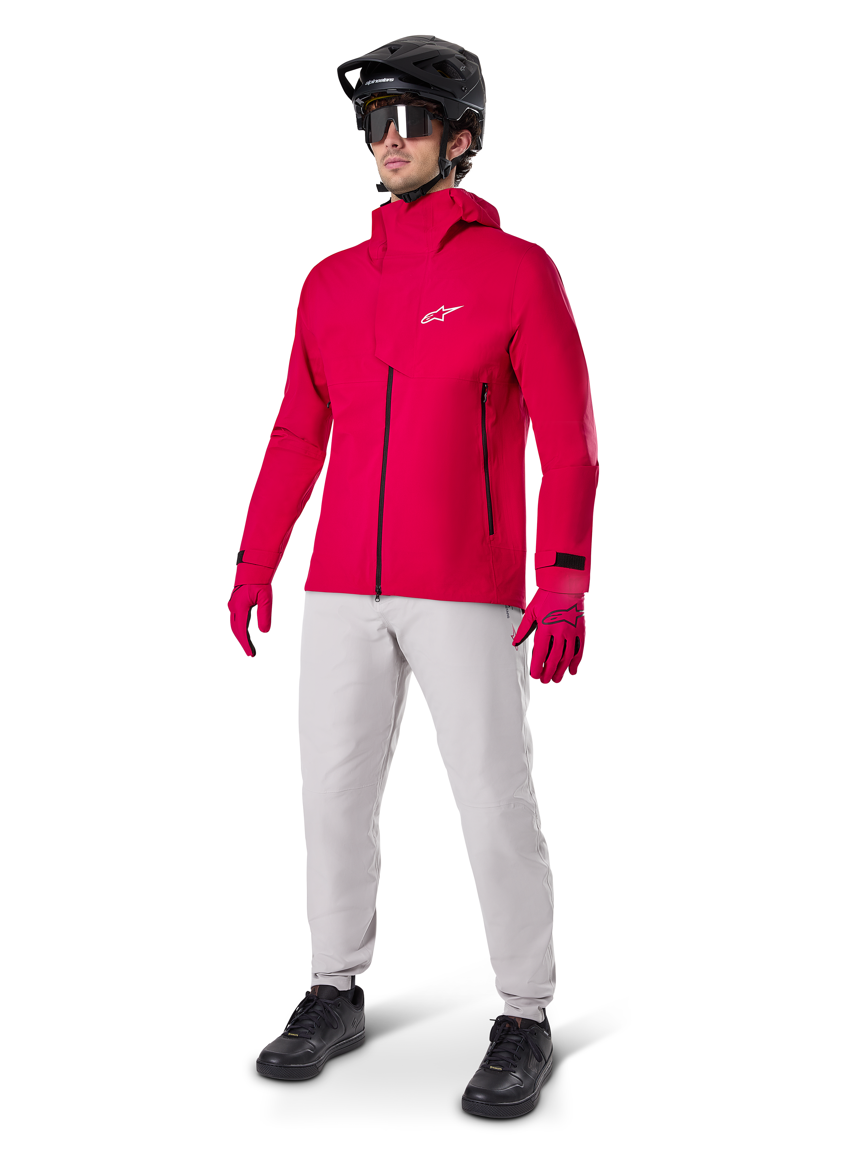 Männlicher Radfahrer in der Alpinestars A-Dura Elite wasserdichten Jacke in Red Fluo, kombiniert mit hellgrauer technischer Hose, roten Handschuhen, schwarzem MTB-Helm mit Brille und schwarzen Schuhen, komplettes Hochleistungs-Outfit für Mountainbiking