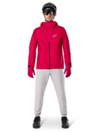 Alpinestars A-Dura Elite Wasserdichte Jacke, Red Fluo, technische Mountainbike-Jacke getragen mit einem schwarzen MTB-Helm, Sonnenbrille, passenden roten Handschuhen und hellgrauer Hose, Ganzkörperansicht einer Allwetter-Fahrradausrüstung