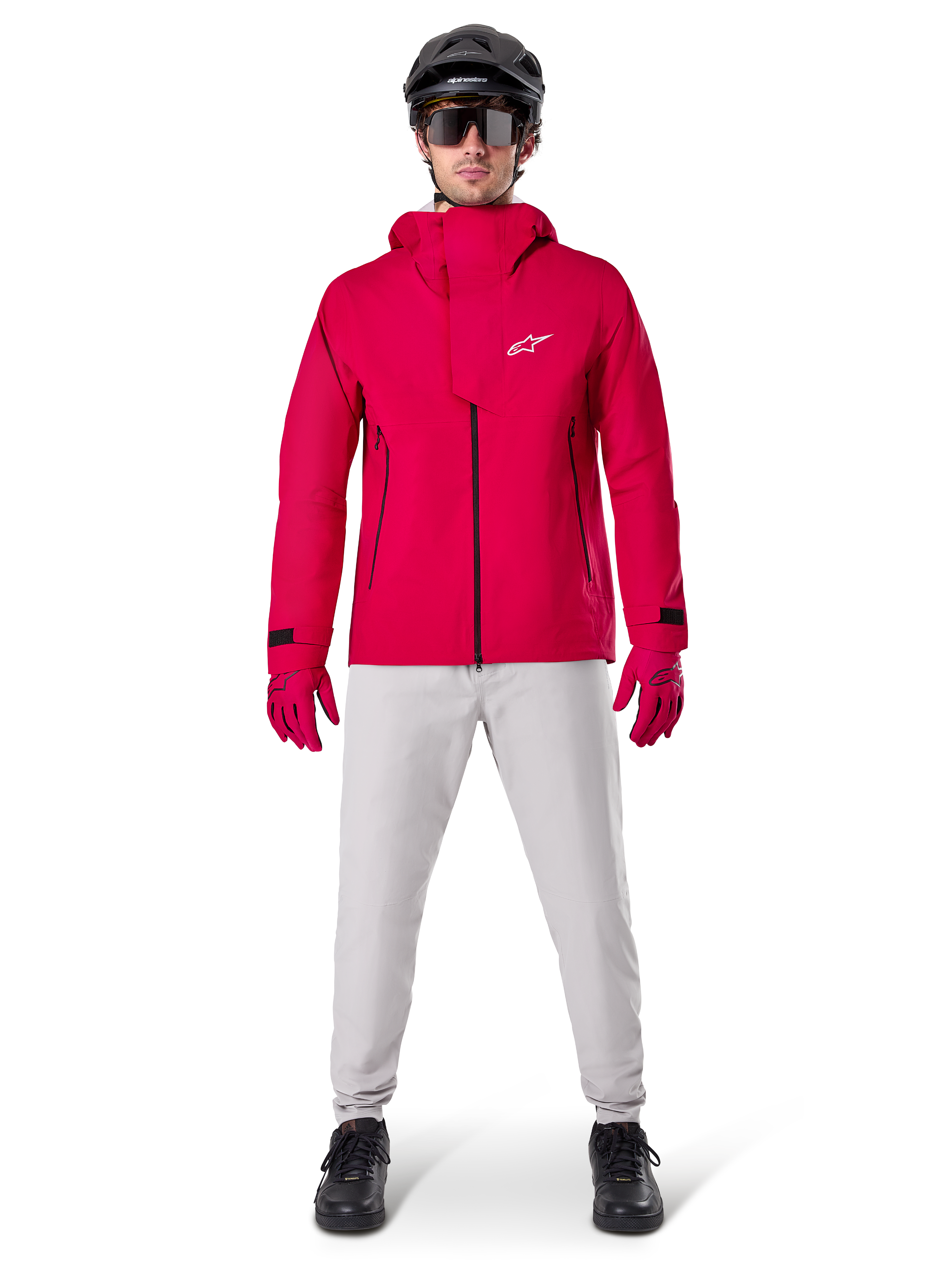 Alpinestars A-Dura Elite Wasserdichte Jacke, Red Fluo, technische Mountainbike-Jacke getragen mit einem schwarzen MTB-Helm, Sonnenbrille, passenden roten Handschuhen und hellgrauer Hose, Ganzkörperansicht einer Allwetter-Fahrradausrüstung