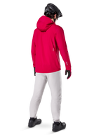 Alpinestars A-Dura Elite Wasserdichte Jacke, Red Fluo, Dreiviertel-Rückansicht, getragen mit hellgrauer Hose und schwarzem MTB-Helm, verfügt über eine Kapuze und reflektierende Akzente, Hochleistungs-Fahrrad-Regenjacke für Trailfahrten