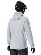 Alpinestars A-Dura Thermojacke, Titanium-Grau, Rückansicht eines Fahrers in der isolierten Kapuzenjacke, kombiniert mit einem schwarzen MTB-Helm und schwarzen Handschuhen, kleines Alpinestars-Logo am unteren Rücken, konzipiert für Mountainbiking bei kaltem Wetter