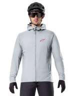Alpinestars A-Dura Thermal Jacket in Titanium, hellgraue Fahrradjacke mit Kapuze und rotem Stern-Logo auf der Brust, getragen von einem Fahrer mit schwarzem MTB-Helm, Sonnenbrille und Handschuhen, isolierte Ausrüstung fürs Mountainbiken