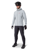 Alpinestars A-Dura Thermojacke in Titanium, hellgraue Fahrradjacke mit Kapuze, rotes Alpinestars-Logo auf der Brust, getragen von einem Mann mit schwarzem MTB-Helm, Sonnenbrille, dunkelgrauer Hose und schwarzen Handschuhen, professionelle Mountainbike-Ausrüstung