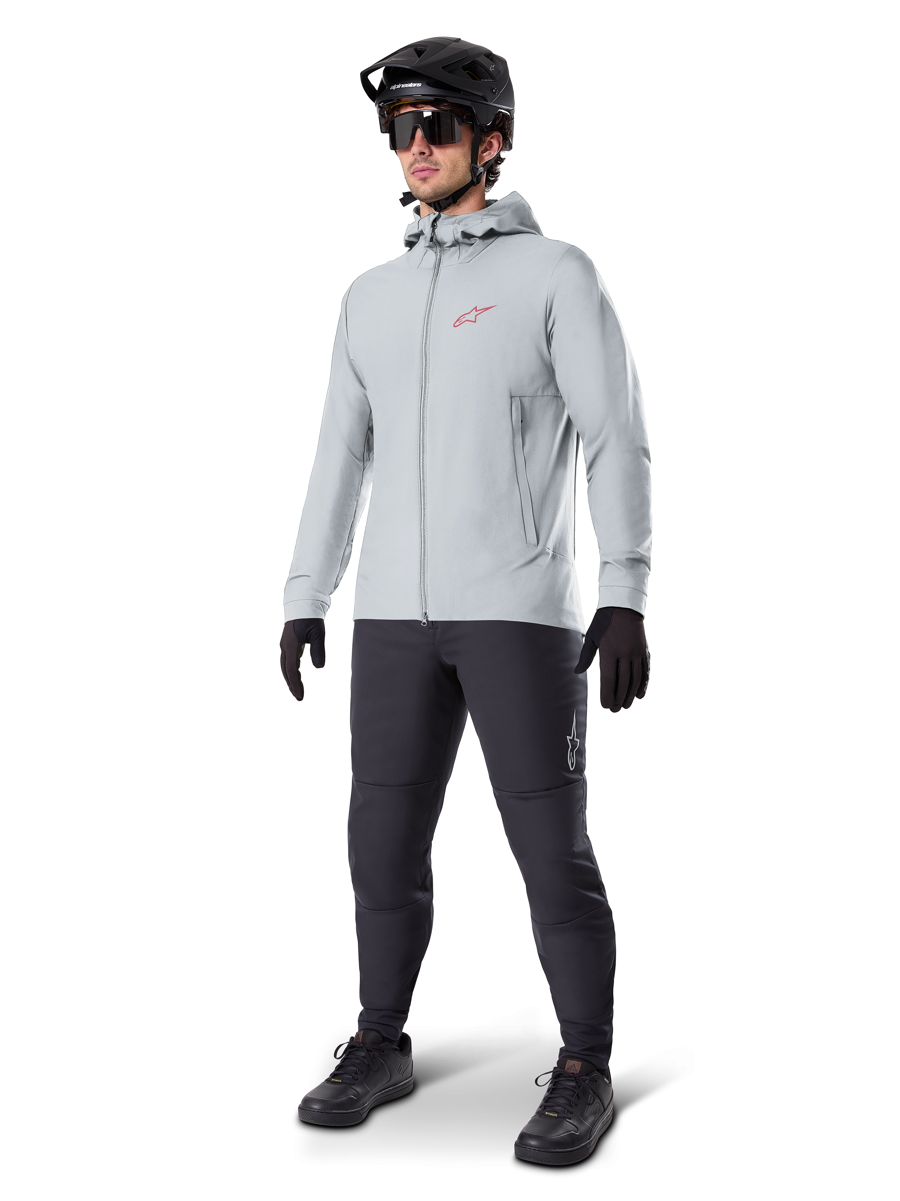 Alpinestars A-Dura Thermojacke in Titanium, hellgraue Fahrradjacke mit Kapuze, rotes Alpinestars-Logo auf der Brust, getragen von einem Mann mit schwarzem MTB-Helm, Sonnenbrille, dunkelgrauer Hose und schwarzen Handschuhen, professionelle Mountainbike-Ausrüstung