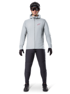 Alpinestars A-Dura Thermojacke, Titanium-Grau, Fahrradjacke getragen mit schwarzer Mountainbike-Hose, Handschuhen und Helm, mit einem kleinen roten Stern-Logo auf der Brust und einem durchgehenden Frontreißverschluss, leichte isolierte Ausrüstung für Offroad-Einsätze.