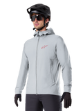 Alpinestars A-Dura Thermojacke, hellgraue isolierte Fahrradjacke, Kapuzendesign mit kleinem rotem Logo, getragen mit schwarzem Helm und Handschuhen, technische Mountainbike-Bekleidung für Fahrten bei kaltem Wetter