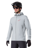 Alpinestars A-Dura Thermojacke, hellgraue isolierte Fahrradjacke, Kapuzendesign mit kleinem rotem Logo, getragen mit schwarzem Helm und Handschuhen, technische Mountainbike-Bekleidung für Fahrten bei kaltem Wetter