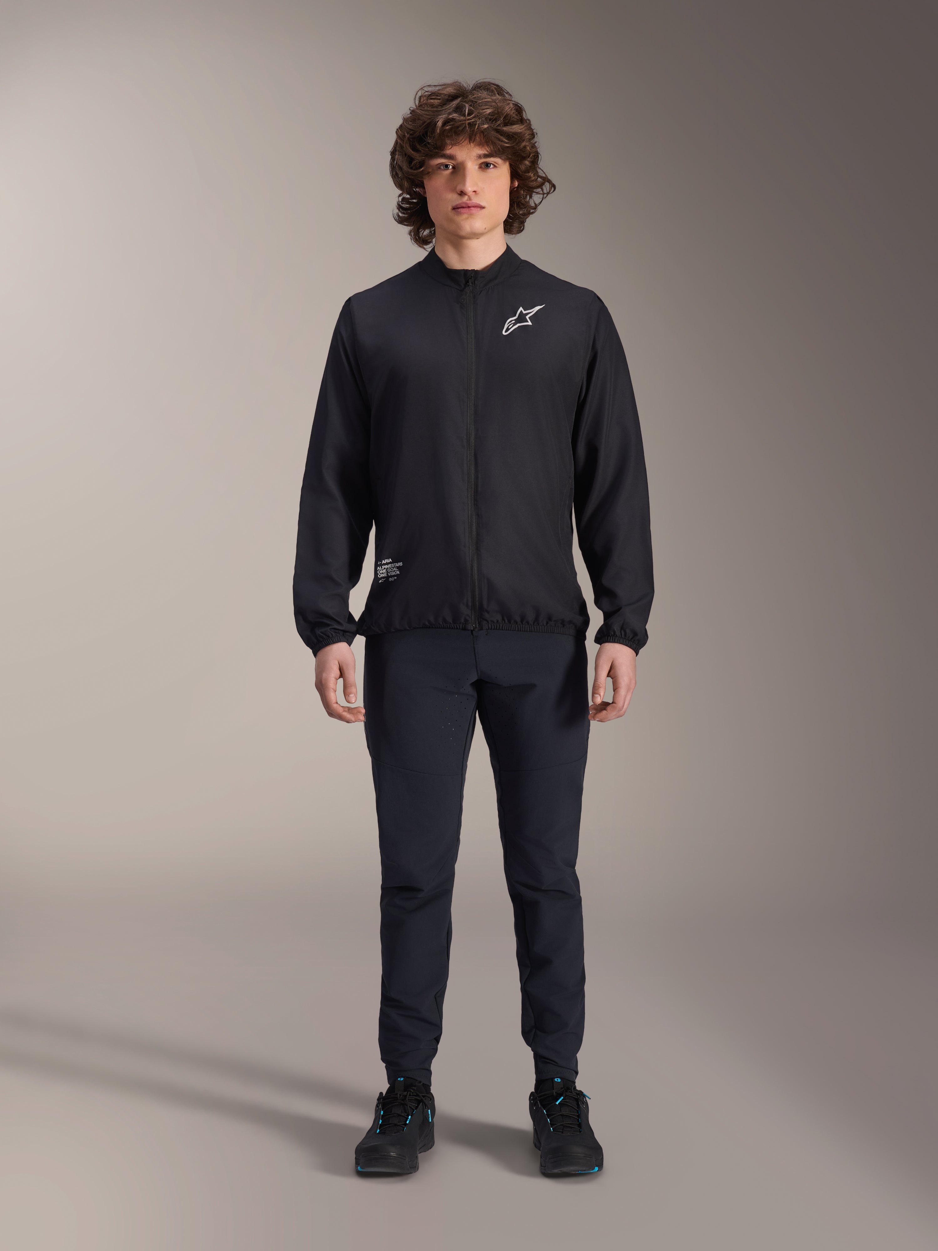 Model trägt die Alpinestars A-Aria Windbreaker-Jacke in Schwarz, mit durchgehendem Front-Reißverschluss, Stehkragen und weißem Astars-Logo auf der Brust, kombiniert mit schwarzer Funktionshose und schwarzen Schuhen, Performance-Radbekleidung für Mountainbiking und Outdoor-Aktivitäten.