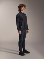 Alpinestars A-Aria Windbreaker Jacke, Schwarz, leichte windabweisende Radjacke, getragen von einem Model mit lockigem Haar, weißes Astars-Logo auf der Brust und Branding unten rechts, kombiniert mit schwarzer Hose und Schuhen, Performance-Passform für Mountainbiking und Gravel-Riding