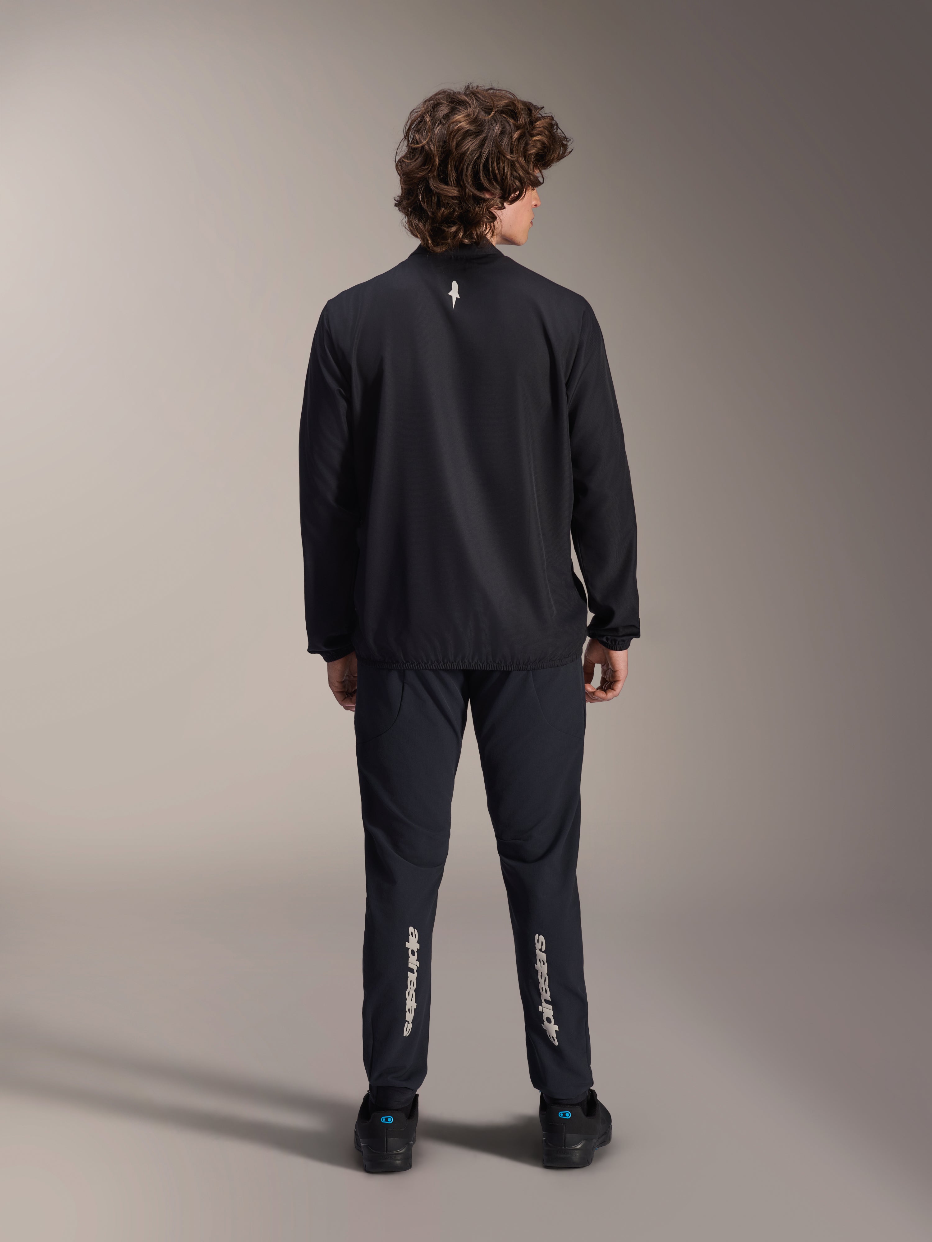 Alpinestars A-Aria Windbreaker-Jacke, Schwarz, Rückansicht, leichte windabweisende Radsportjacke mit einem kleinen weißen Logo auf dem oberen Rücken, getragen mit passender schwarzer Hose mit vertikalem Alpinestars-Branding an den Waden, Performance-Mountainbike-Ausrüstung