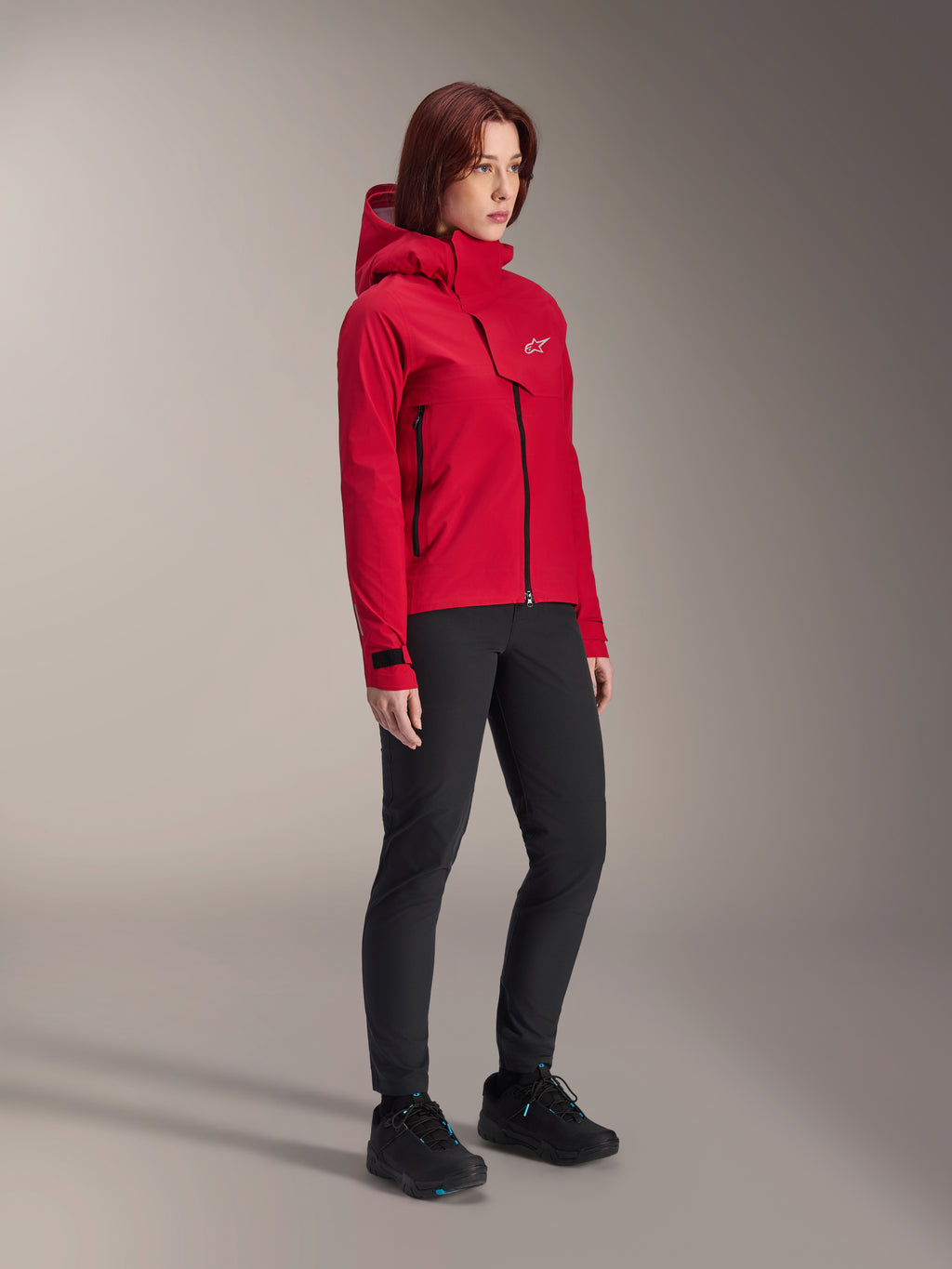 Alpinestars Stella A-Dura Elite wasserdichte Damenjacke, Red Fluo, Mountainbike-Regenjacke mit Kapuze und weißem Astars-Logo auf der Brustklappe, getragen mit schwarzer Hose und schwarzen Mountainbike-Schuhen, technische Performance-Ausrüstung für Trailfahrten