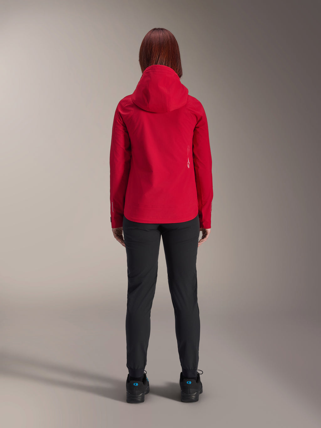 Rückansicht einer Frau, die die Alpinestars Stella A-Dura Elite Wasserdichte Jacke in Red Fluo trägt, kombiniert mit einer schwarzen Hose und Schuhen, ausgestattet mit einer schützenden Kapuze und einem verlängerten Rückensaum für das Mountainbiken.