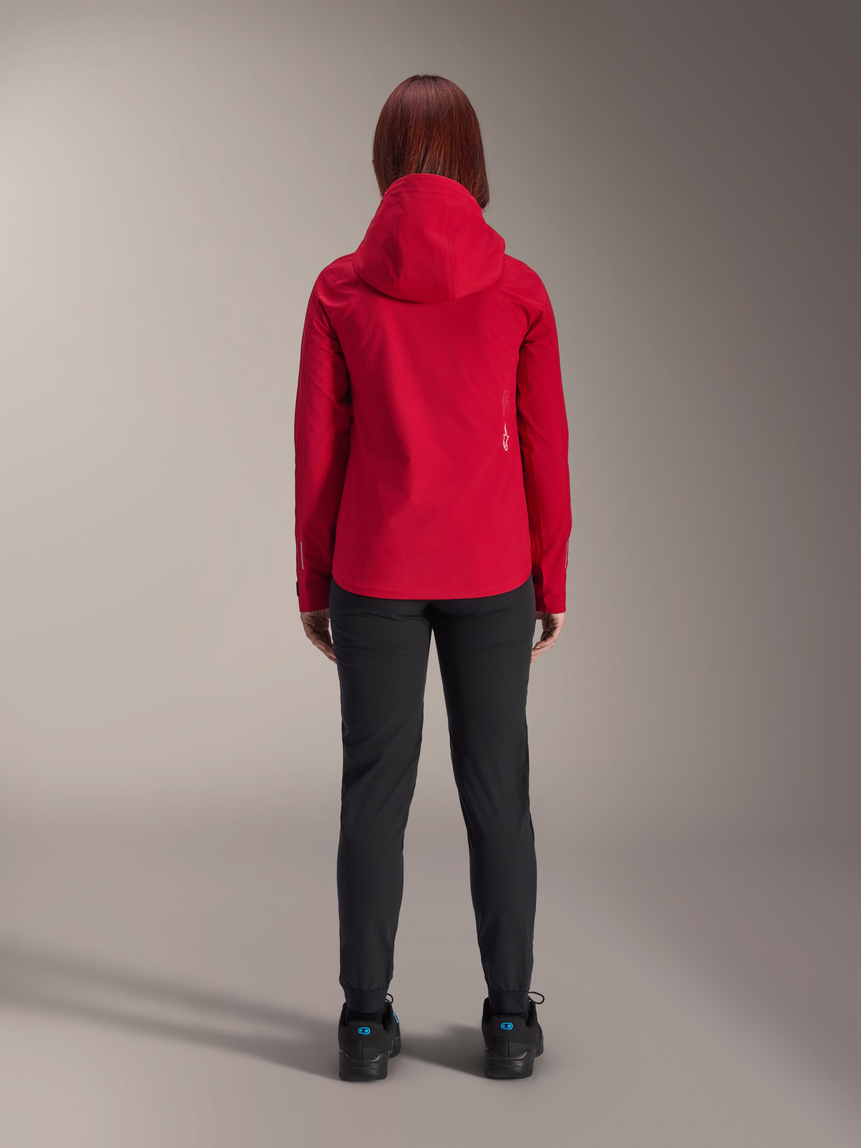 Rückansicht einer Frau, die die Alpinestars Stella A-Dura Elite Wasserdichte Jacke in Red Fluo trägt, kombiniert mit einer schwarzen Hose und Schuhen, ausgestattet mit einer schützenden Kapuze und einem verlängerten Rückensaum für das Mountainbiken.