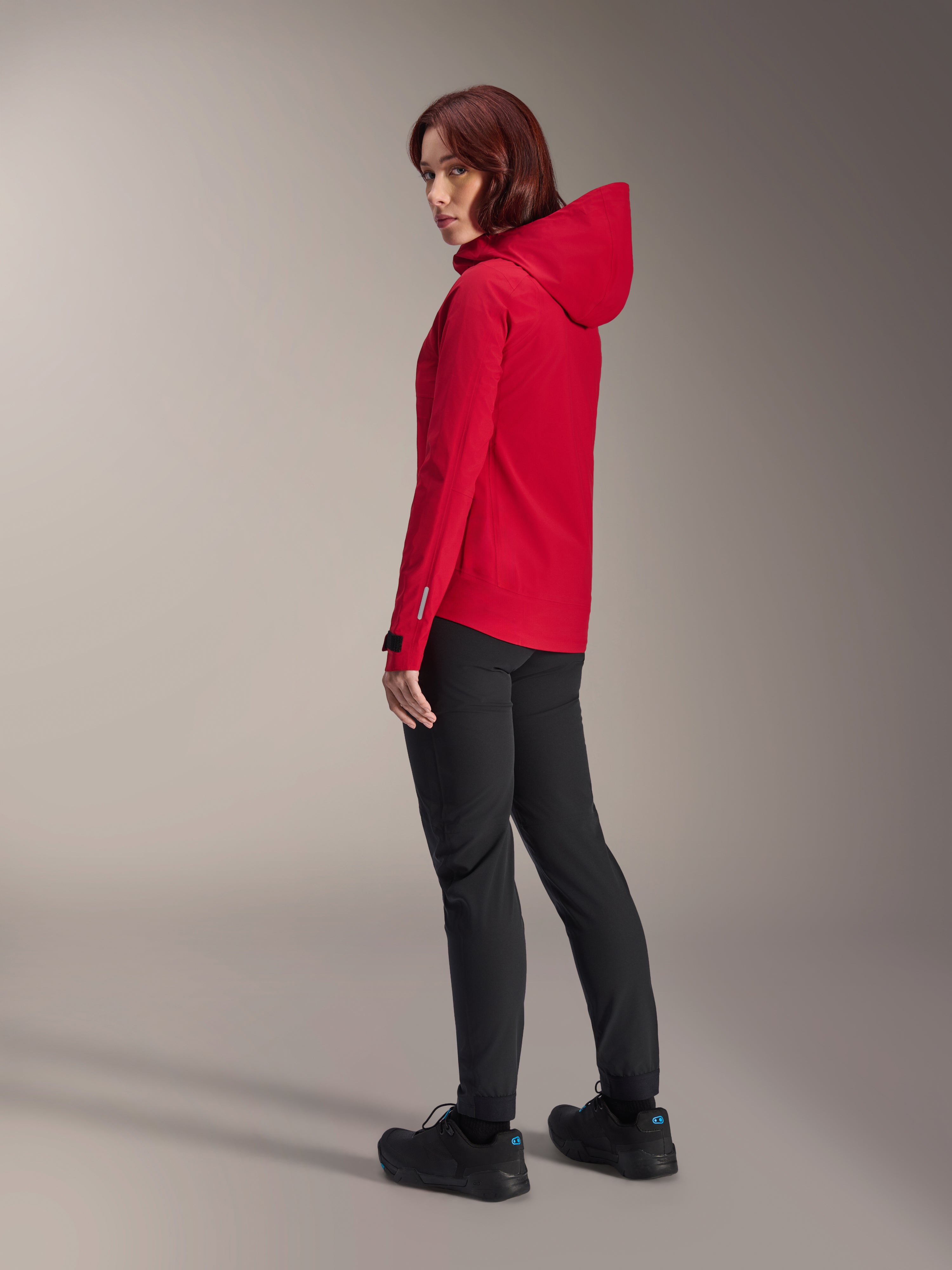 Alpinestars Stella A-Dura Elite Wasserdichte Damenjacke in Red Fluo, kombiniert mit schwarzer technischer Radhose und schwarzen Schuhen, Dreiviertel-Rückansicht mit verstellbarer Kapuze und verlängertem Rückensaum, Performance-Mountainbike-Bekleidung.