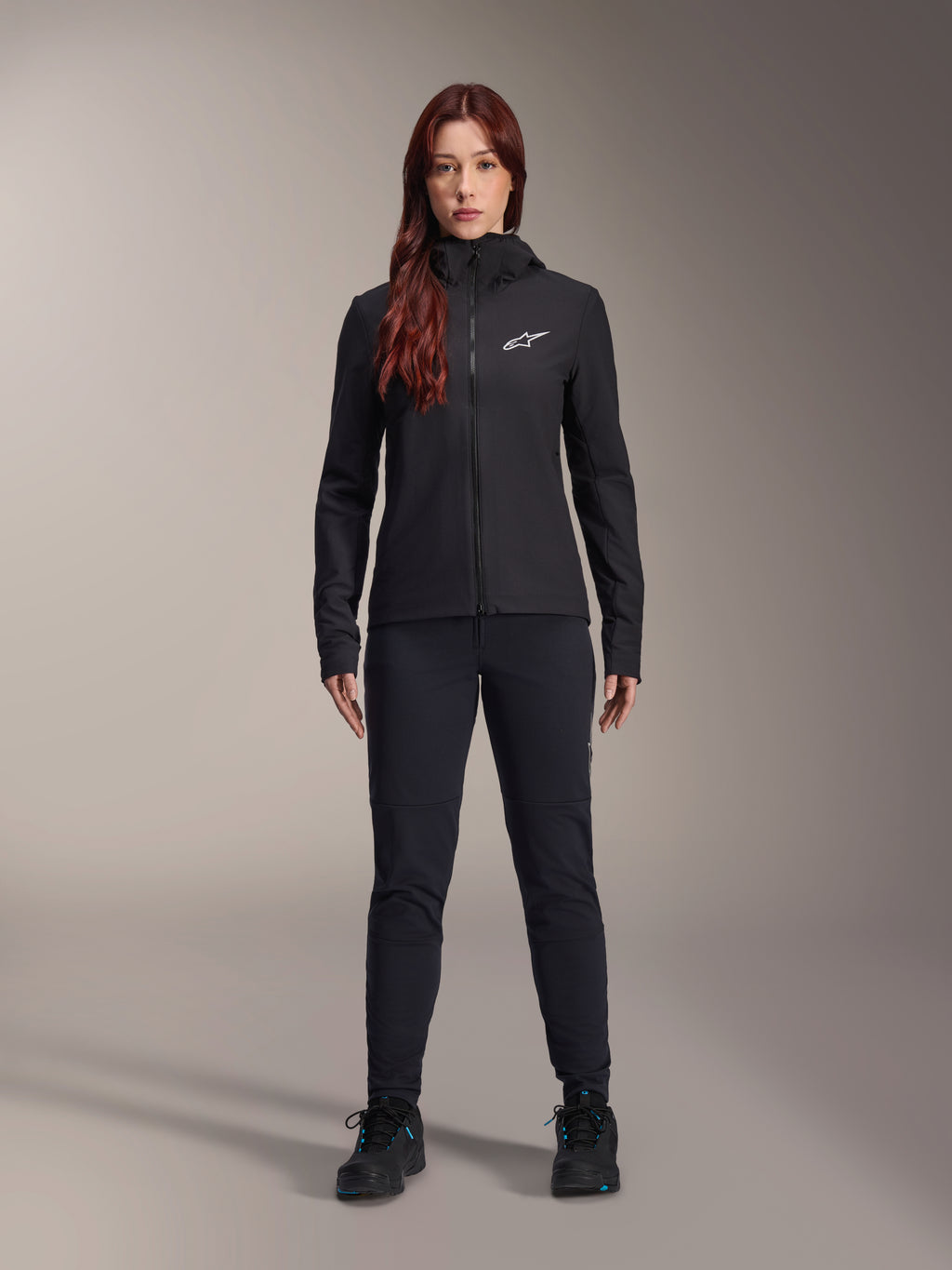 Frau trägt die Alpinestars Stella A-Dura Thermal Jacke, Schwarz, leichte isolierte Fahrradjacke mit Kapuze und weißem Astars-Logo auf der Brust, kombiniert mit schwarzer Hose und Sportschuhen