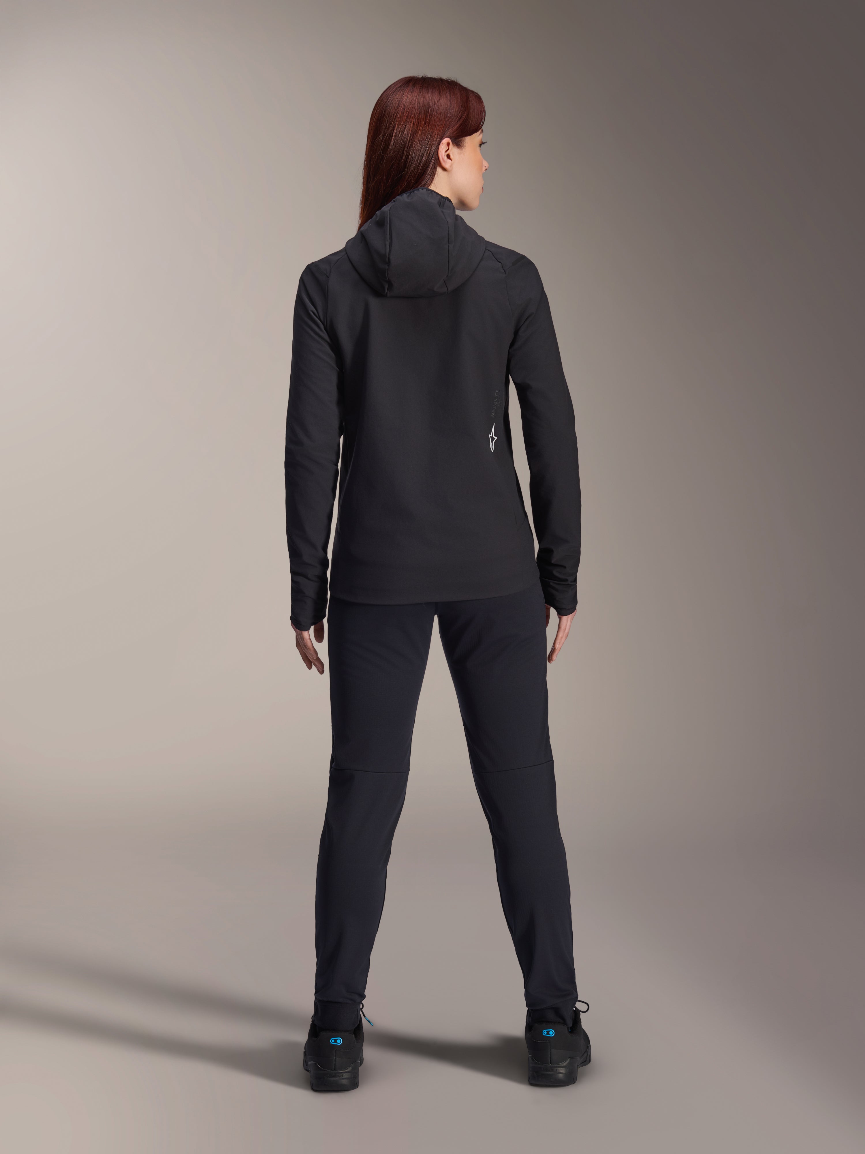 Alpinestars Stella A-Dura Thermal Jacke für Damen, Rückansicht, schwarze isolierte Fahrradjacke mit Kapuze, passende schwarze Hose und Schuhe, kleines weißes Logo auf dem unteren Rücken