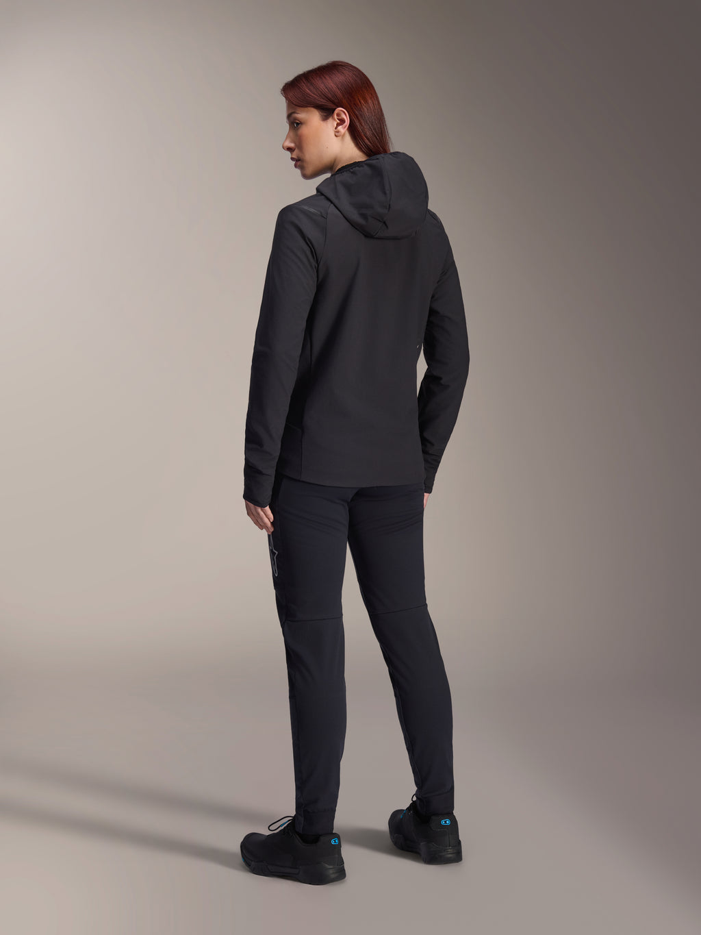 Alpinestars Stella A-Dura Thermal Jacket, MTB-Fahrradjacke für Damen, Schwarz, Rückansicht mit Fokus auf die isolierte Kapuze und die reguläre Passform, getragen mit passender schwarzer Hose und Schuhen, leichte Wärmeisolierung für Fahrten bei kaltem Wetter