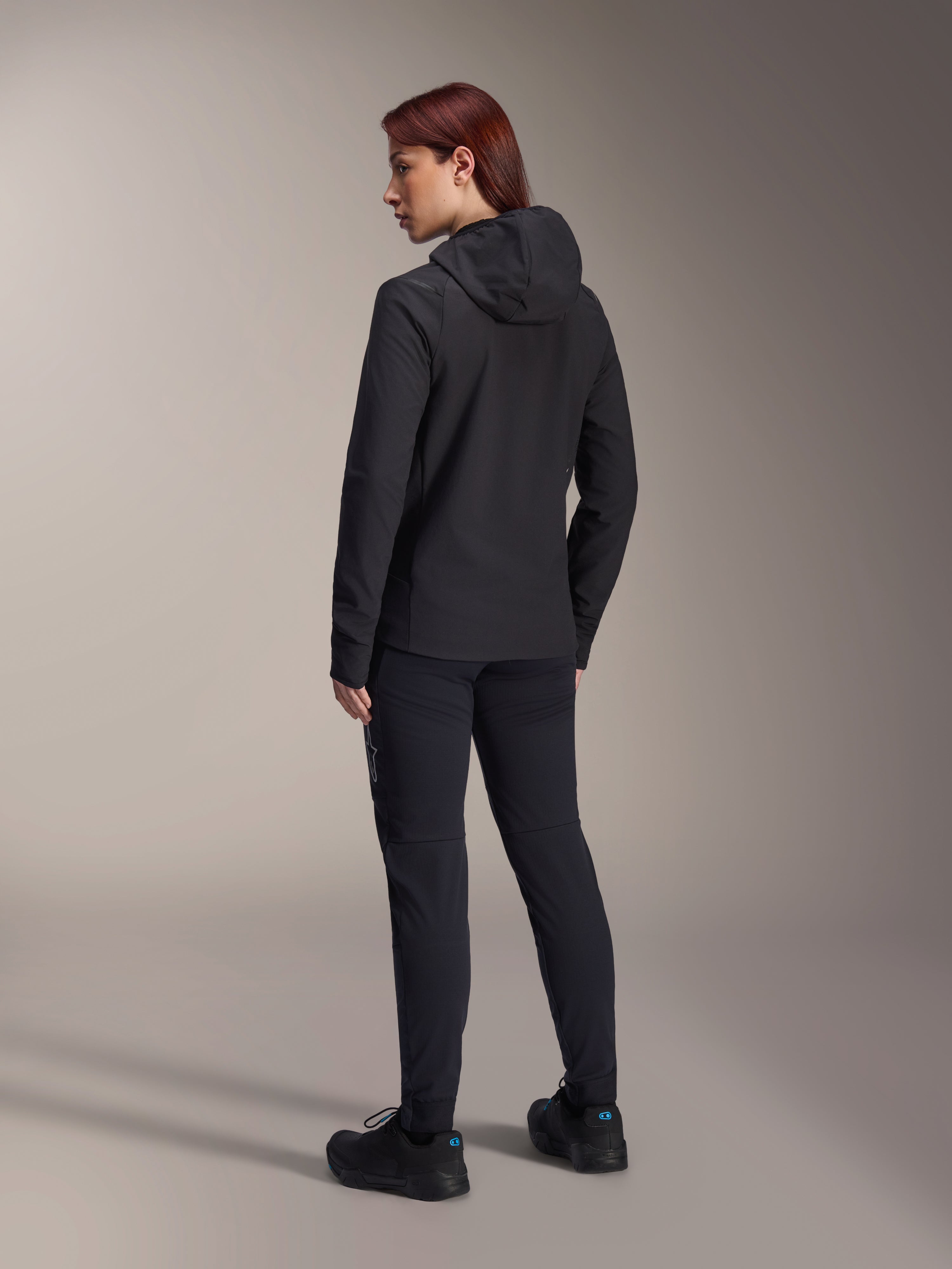 Alpinestars Stella A-Dura Thermal Jacket, MTB-Fahrradjacke für Damen, Schwarz, Rückansicht mit Fokus auf die isolierte Kapuze und die reguläre Passform, getragen mit passender schwarzer Hose und Schuhen, leichte Wärmeisolierung für Fahrten bei kaltem Wetter