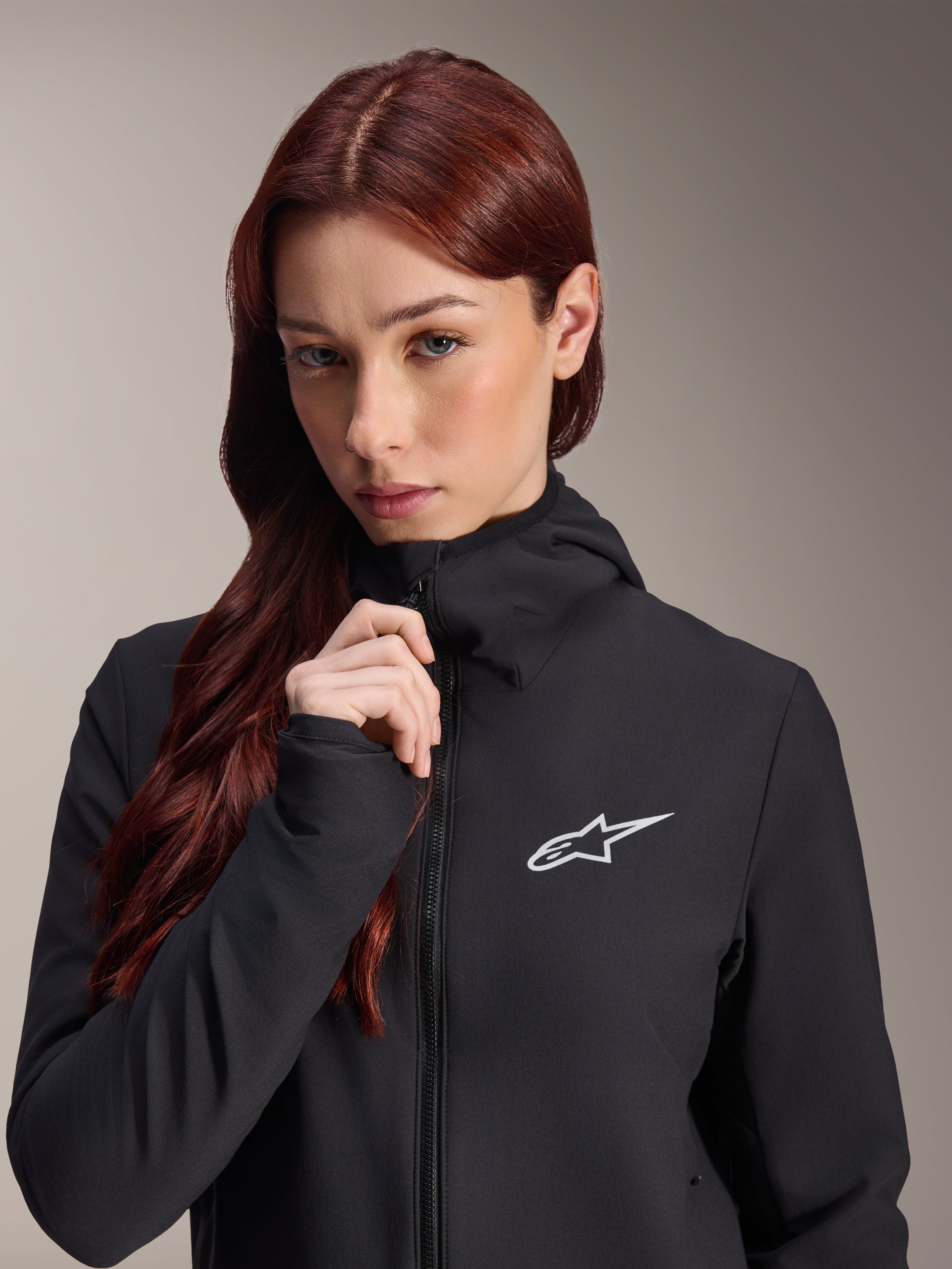 Alpinestars Stella A-Dura Thermal Jacke, schwarze Damen-Fahrradjacke, getragen von einem Model mit langem rötlich-braunem Haar, weißes Alpinestars-Logo auf der Brust, Kapuzendesign mit durchgehendem Reißverschluss, Mountainbike-Bekleidung