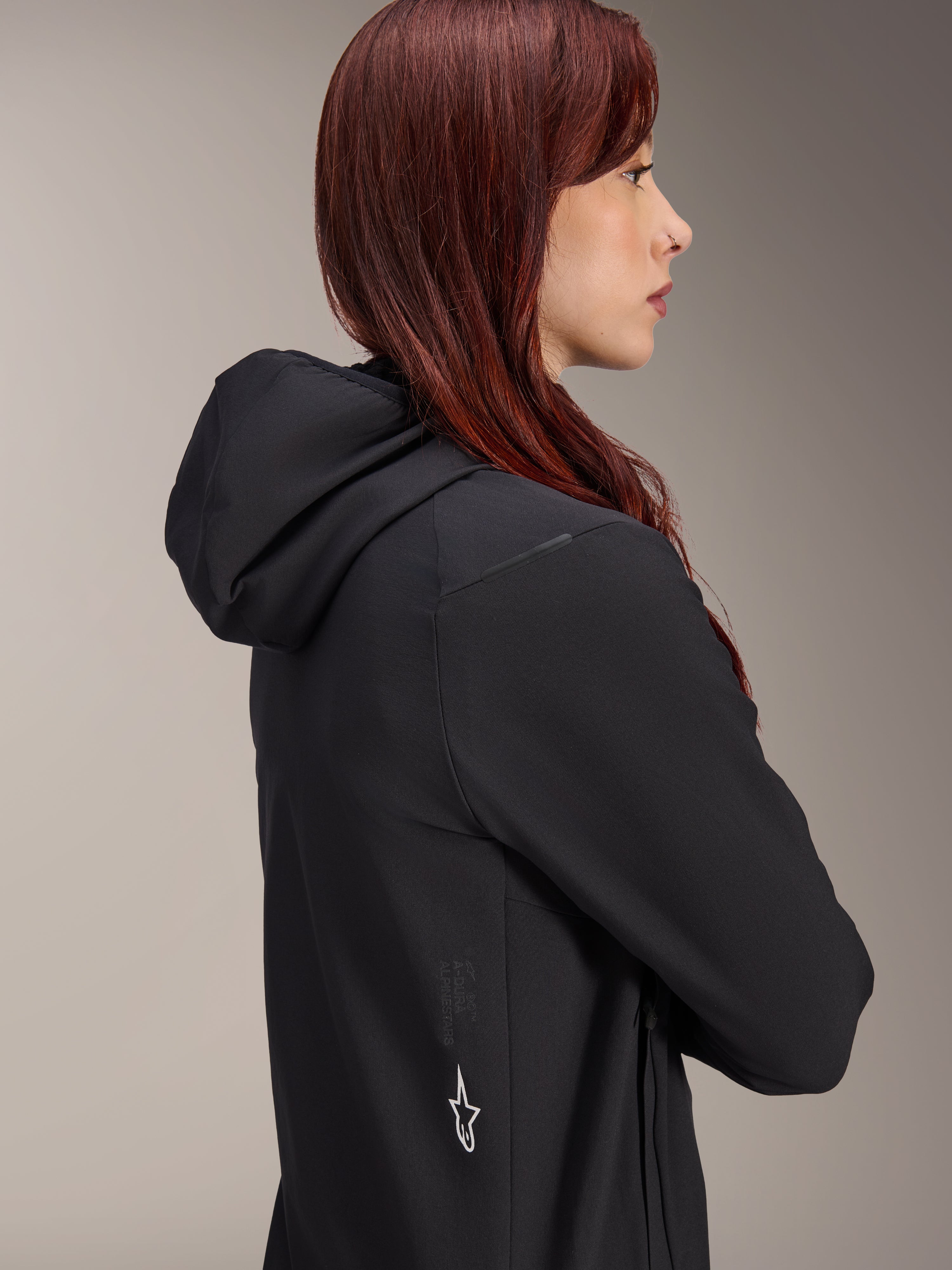 Alpinestars Stella A-Dura Thermojacke, Damen-Fahrradjacke, Schwarz, Seitenprofil einer Frau mit roten Haaren, die die Jacke trägt, verfügt über eine integrierte Kapuze und ein weißes Alpinestars Stern-Logo an der Seite, leichte isolierte Thermo-Ausrüstung für MTB.