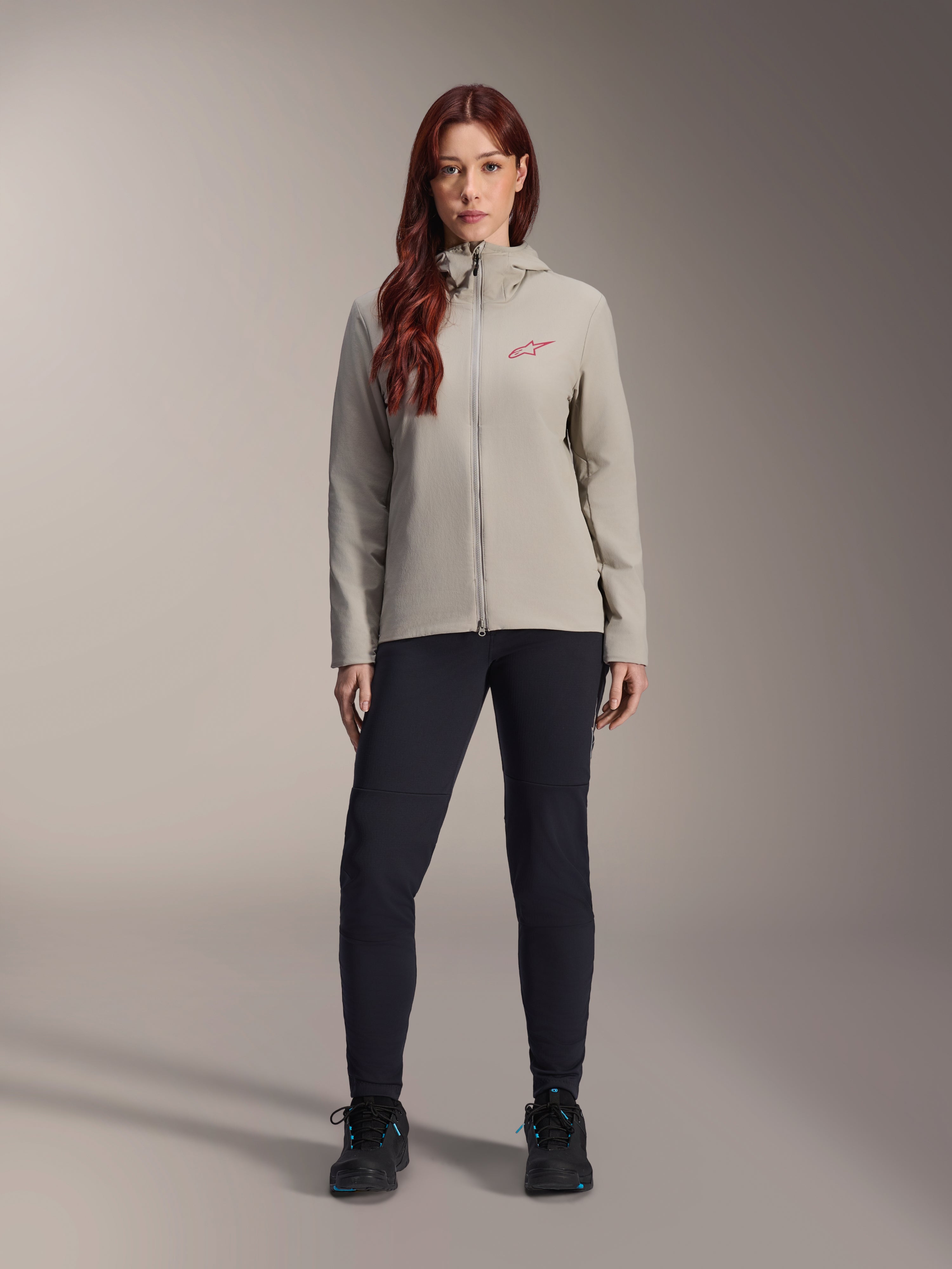 Alpinestars Stella A-Dura Thermal Damenjacke, Mountainbike-Jacke, Titanium-Grau, getragen mit schwarzer Funktionshose und schwarzen Schuhen, mit fleecegefütterter Kapuze und rotem Logo auf der Brust, konzipiert für Fahrten bei kaltem Wetter