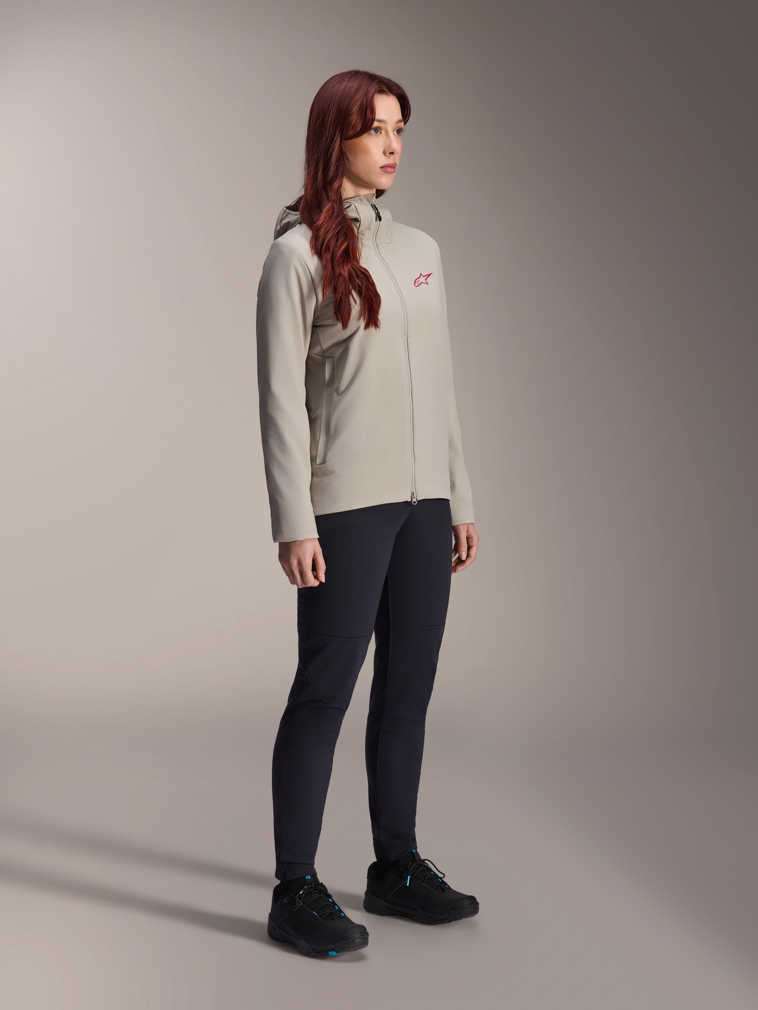 Alpinestars Stella A-Dura Thermal Jacke, Titanium-Grau, Damen-Thermo-Fahrradjacke mit rotem Stern-Logo auf der Brust, getragen mit schwarzer technischer Hose und schwarzen Mountainbike-Schuhen, Studioaufnahme für Outdoor-Radbekleidung