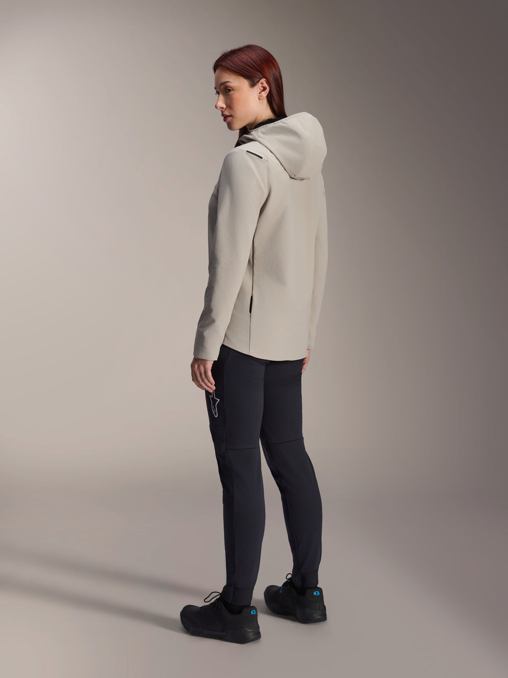 Alpinestars Stella A-Dura Thermal Jacke in Titanium Grau, Dreiviertel-Rückansicht einer Frau, die die isolierte Fahrradjacke mit Kapuze und eine schwarze MTB-Hose trägt, Studioaufnahme zeigt die reguläre Passform und Taschendetails.