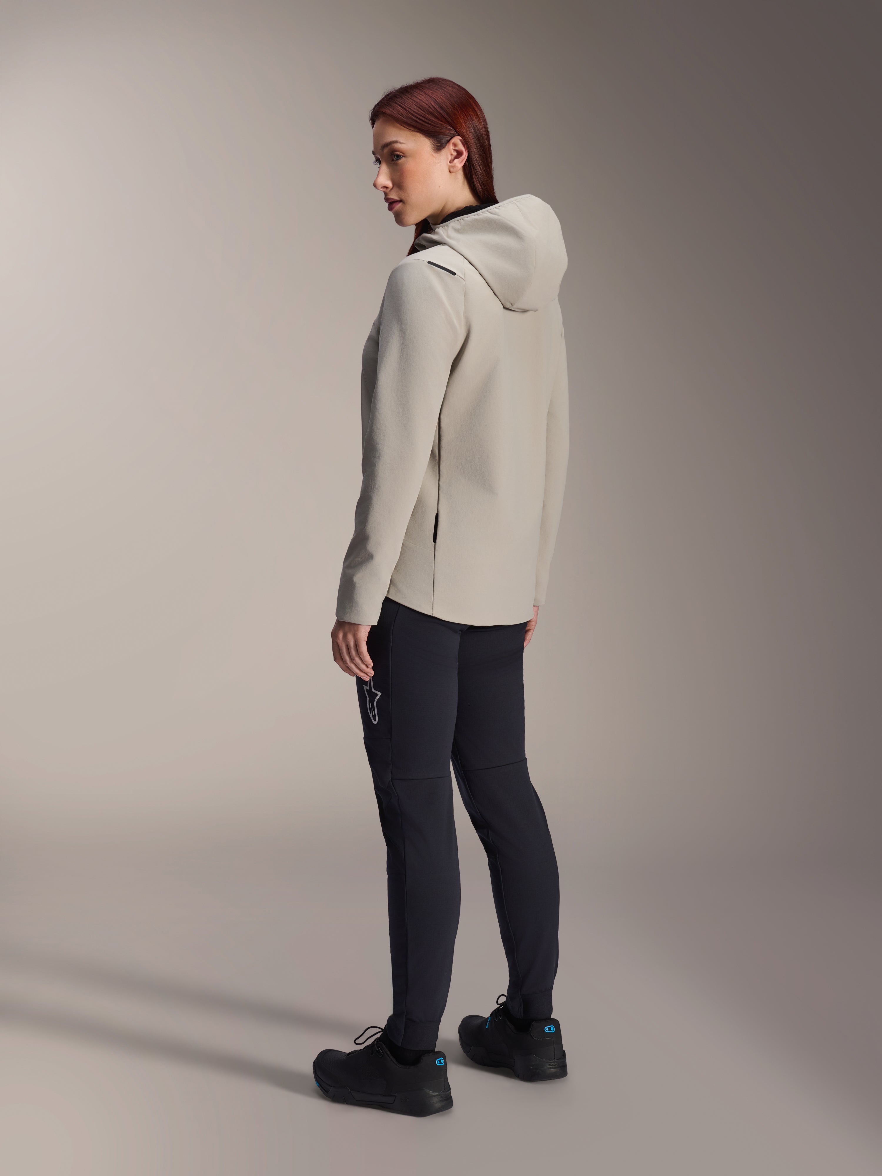 Alpinestars Stella A-Dura Thermal Jacke in Titanium Grau, Dreiviertel-Rückansicht einer Frau, die die isolierte Fahrradjacke mit Kapuze und eine schwarze MTB-Hose trägt, Studioaufnahme zeigt die reguläre Passform und Taschendetails.