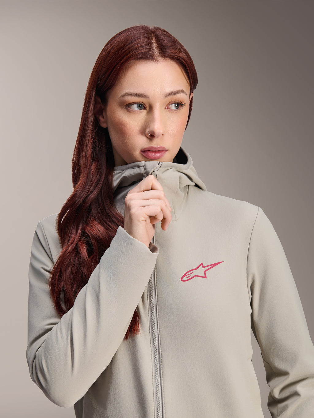 Alpinestars Stella A-Dura Thermojacke für Damen, Titanium-Grau mit pinkfarbenem Alpinestars Stern-Logo, Modell mit langem rotem Haar trägt die Kapuzenjacke und schließt den Reißverschluss, leichte isolierte Radbekleidung für kaltes Wetter