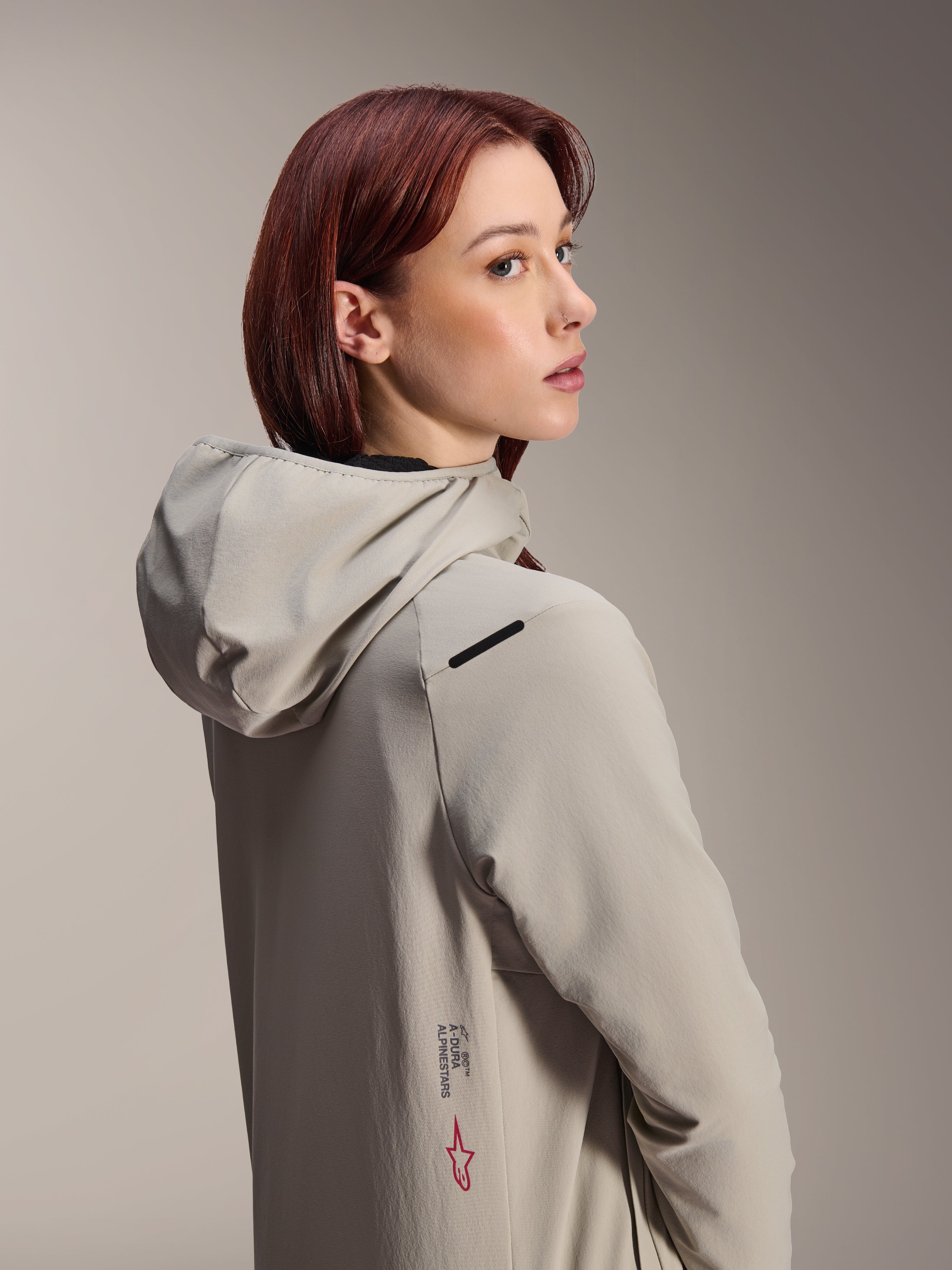 Alpinestars Stella A-Dura Thermal Jacket, Damen-MTB-Jacke, Titanium-Grau, Frau, die über ihre Schulter blickt und eine Performance-Jacke mit Kapuze trägt, mit Alpinestars-Schriftzug und rotem Stern-Logo an der Seite, konzipiert für Radsport bei kaltem Wetter