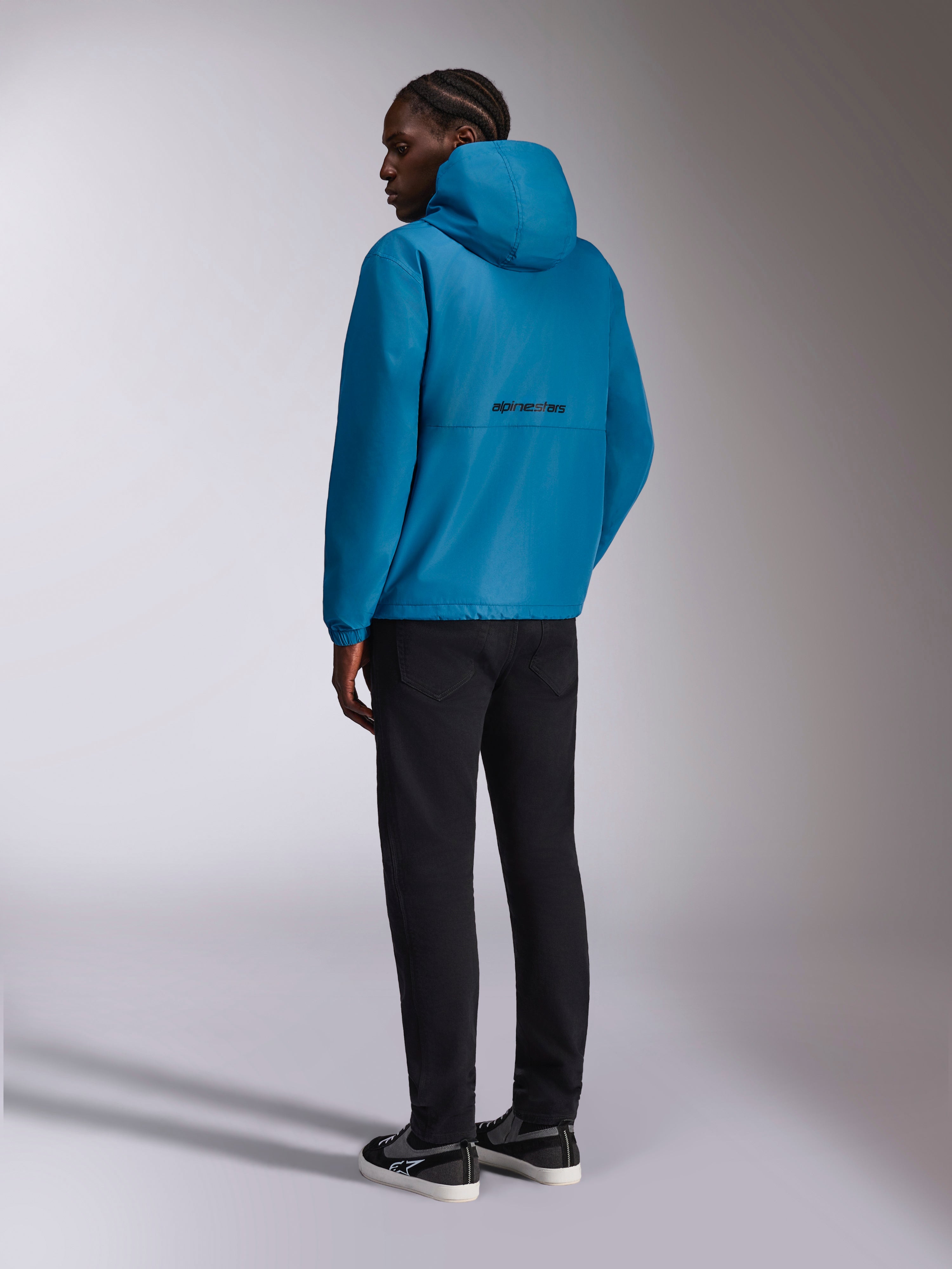 Alpinestars Speeder Chromium Windbreaker-Jacke, Kapuzen-Windjacke, Blau, Rückansicht mit schwarzem Alpinestars-Wortlogo auf dem Rücken, getragen mit schwarzer Hose und schwarz-weißen Sneakern, lässige Motorsport-inspirierte Streetwear
