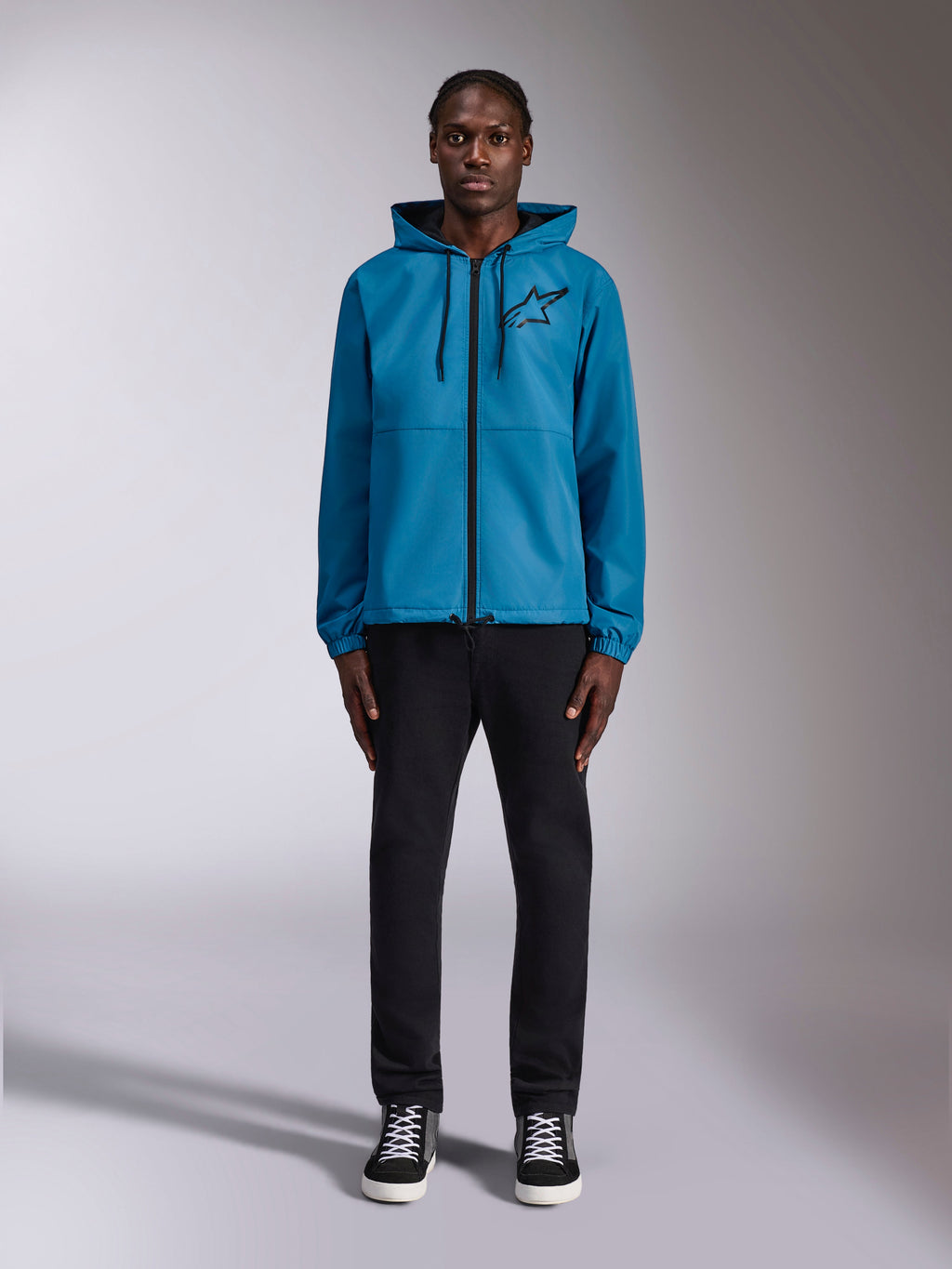 Model trägt die Alpinestars Speeder Chromium Windbreaker-Jacke in Blau, mit Kapuze, schwarzen Kordelzügen, schwarzem Frontreißverschluss und schwarzem Astars-Logo auf der Brust, kombiniert mit einer schwarzen Hose und Sneakern, lässige Motorsport-Streetwear