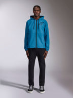 Model trägt die Alpinestars Speeder Chromium Windbreaker-Jacke in Blau, mit Kapuze, schwarzen Kordelzügen, schwarzem Frontreißverschluss und schwarzem Astars-Logo auf der Brust, kombiniert mit einer schwarzen Hose und Sneakern, lässige Motorsport-Streetwear