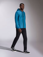 Alpinestars Speeder Chromium Windbreak Jacket, blaue Windjacke mit Kapuze getragen von einem männlichen Model, verfügt über einen schwarzen Frontreißverschluss und ein Brustlogo, kombiniert mit schwarzer Hose und High-Top-Sneakern, lässige vom Motorsport inspirierte Sportbekleidung