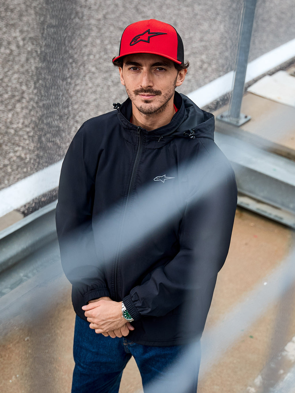 Ein Mann trägt die Alpinestars Overhaul Racer Jacke in Schwarz, eine wasserabweisende Regenjacke mit Kapuze und kleinem Brustlogo, kombiniert mit einer roten Alpinestars-Kappe, lässige Motorsport-Lifestyle-Bekleidung