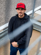 Ein Mann trägt die Alpinestars Overhaul Racer Jacke in Schwarz, eine wasserabweisende Regenjacke mit Kapuze und kleinem Brustlogo, kombiniert mit einer roten Alpinestars-Kappe, lässige Motorsport-Lifestyle-Bekleidung