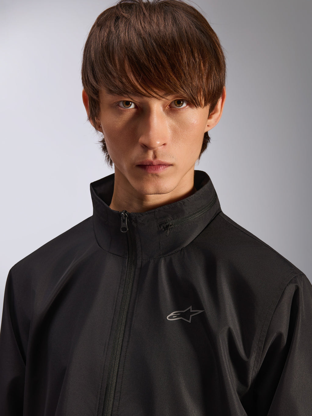 Alpinestars Overhaul Racer Jacke, schwarze Regenjacke, Porträt-Nahaufnahme eines männlichen Models, das die Jacke trägt, mit hohem Kragen und kleinem silbernem Astars-Logo auf der Brust, motorsport-inspirierte Freizeitbekleidung