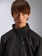 Alpinestars Overhaul Racer Jacke, schwarze Regenjacke, Porträt-Nahaufnahme eines männlichen Models, das die Jacke trägt, mit hohem Kragen und kleinem silbernem Astars-Logo auf der Brust, motorsport-inspirierte Freizeitbekleidung
