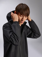 Alpinestars Overhaul Racer Jacke, schwarze Regenjacke mit Kapuze, getragen von einem Model mit aufgesetzter Kapuze, wasserabweisendes 2,5L-Gewebe, elastische Bündchen und minimalistisches Logo auf der Brust, lässige vom Motorsport inspirierte Oberbekleidung