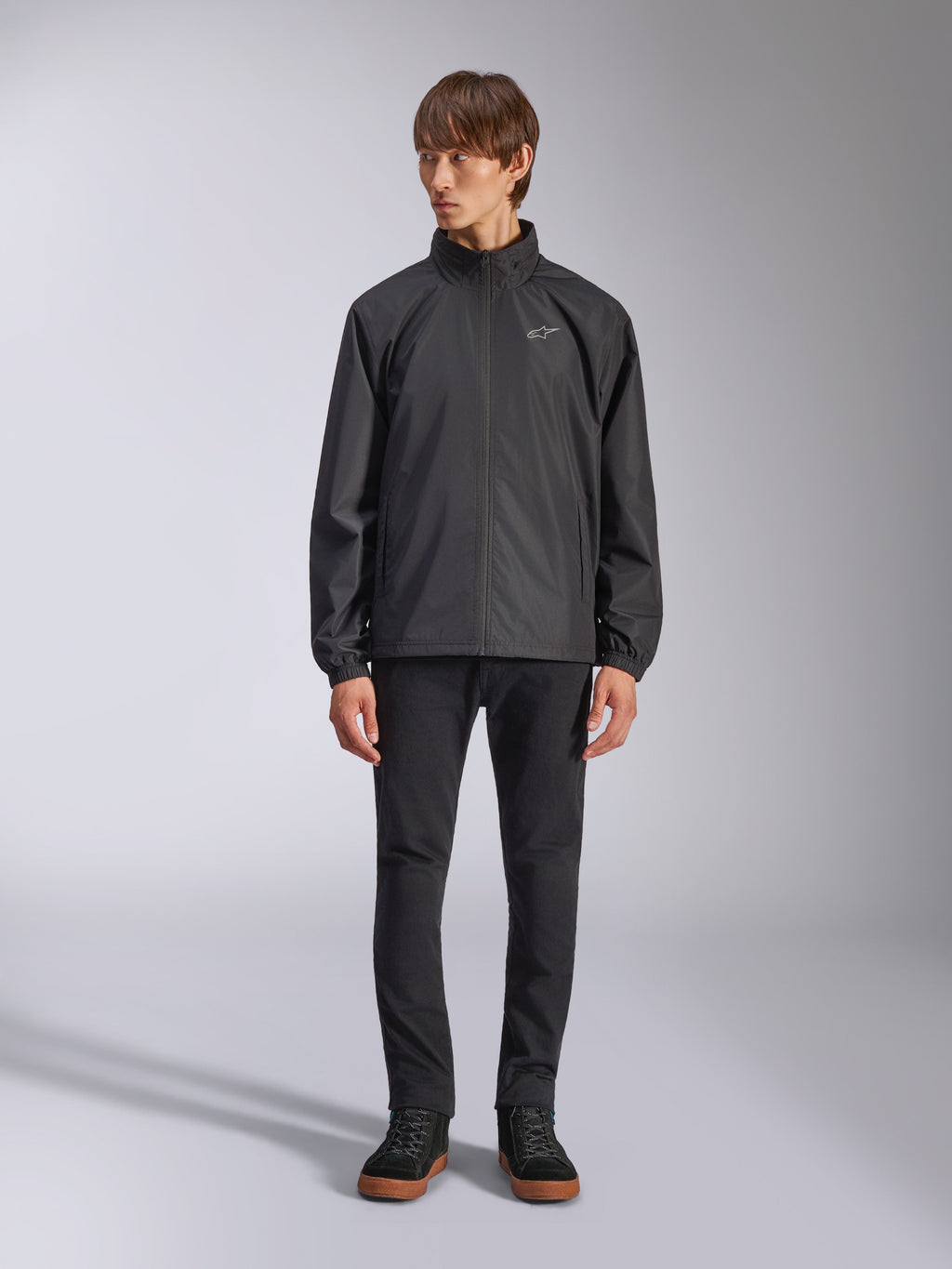 Alpinestars Overhaul Racer Jacke, schwarze Regenjacke für Herren, Ganzkörperansicht an einem Model kombiniert mit schwarzer Hose und lässigen Sneakern, mit kleinem weißen Logo auf der Brust, wasserabweisendem Material und lässigem, vom Motorsport inspiriertem Design